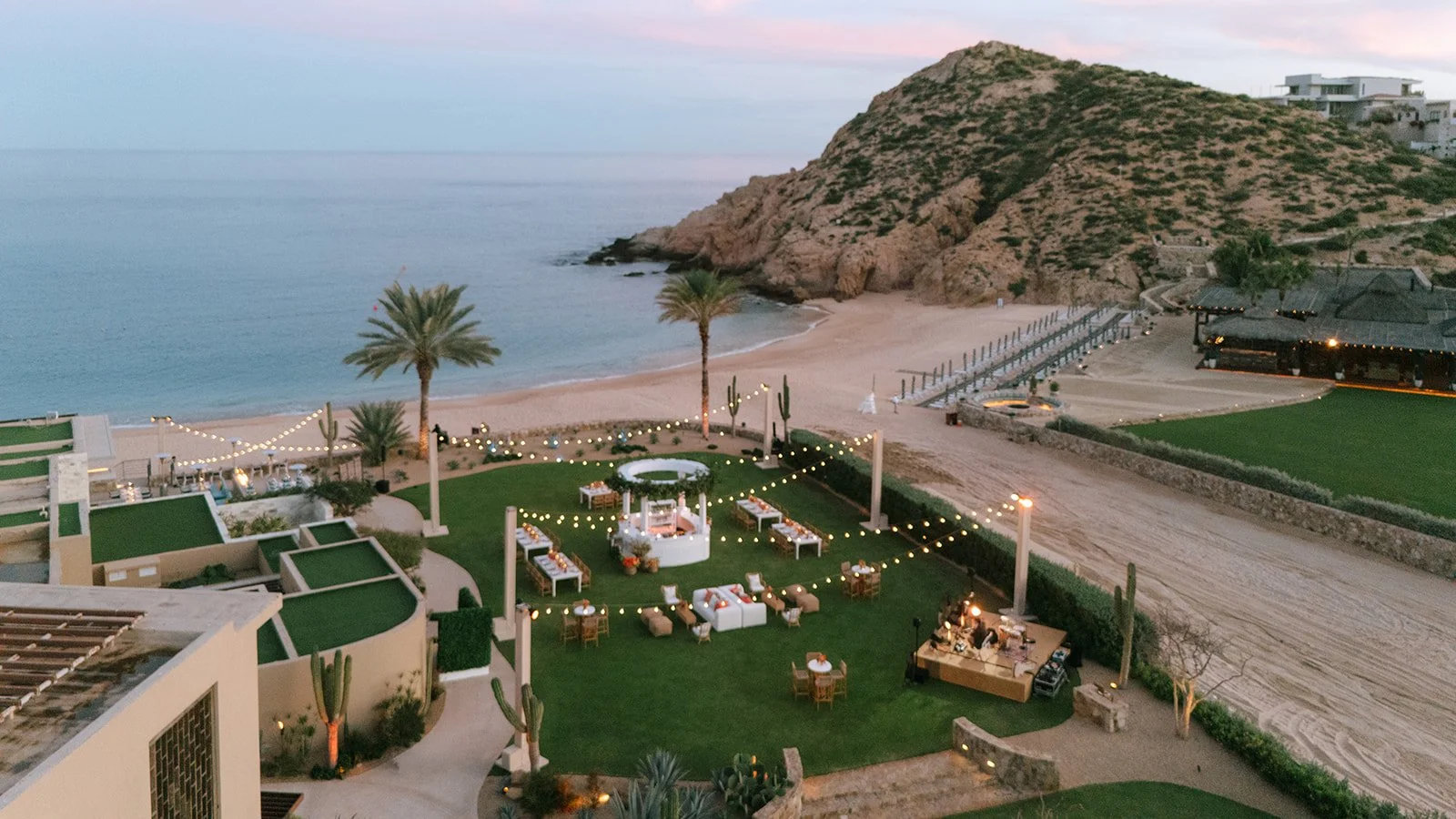 Beachside event space with lounge chairs, tables, string lights, palm trees, and a view of the ocean and rocky hillside at sunset.