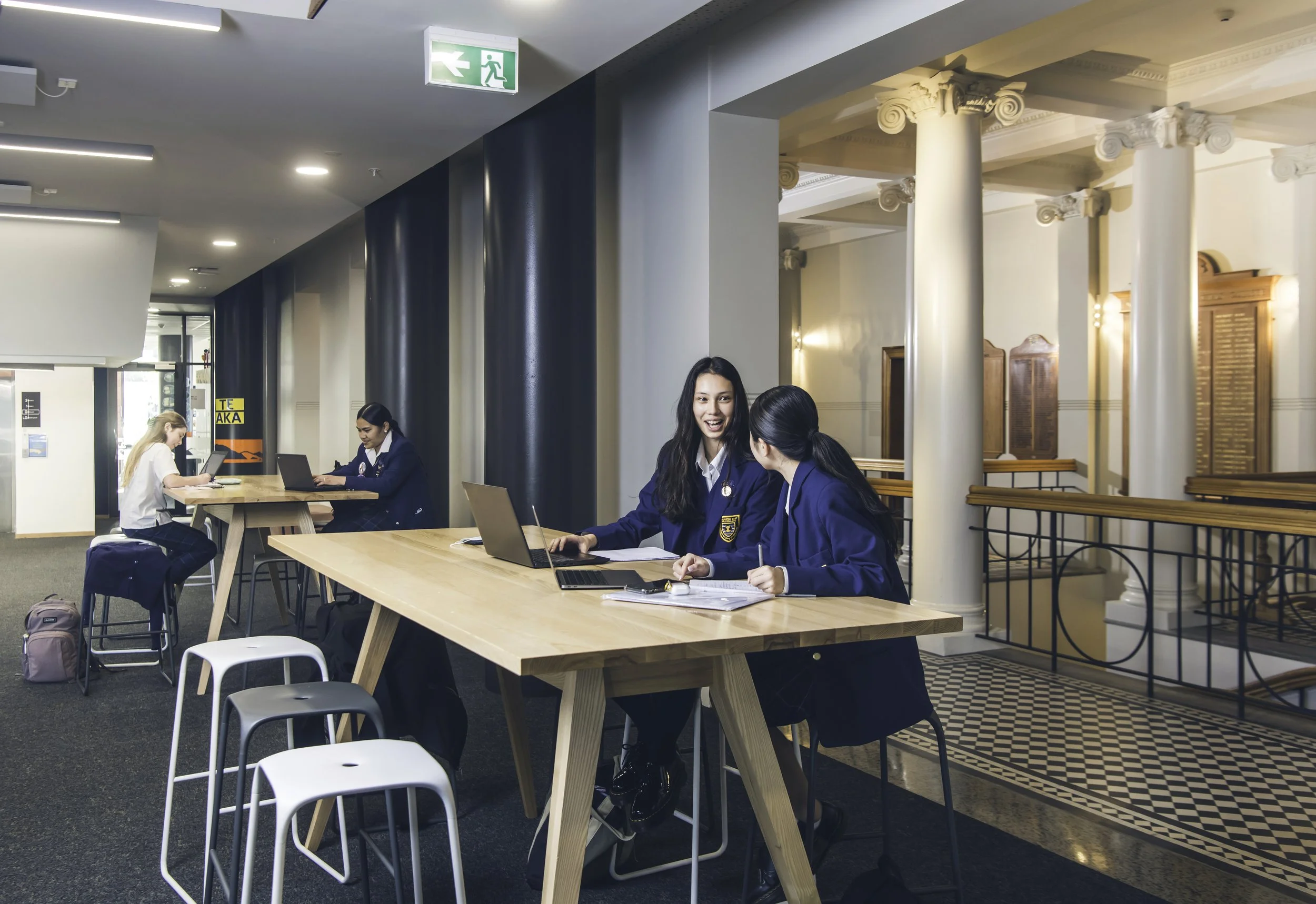 Three students in school uniforms working on laptops and studying at a large wooden table in a modern, well-lit indoor space. Two girls are smiling and chatting, while another girl is focused on her laptop in the background.