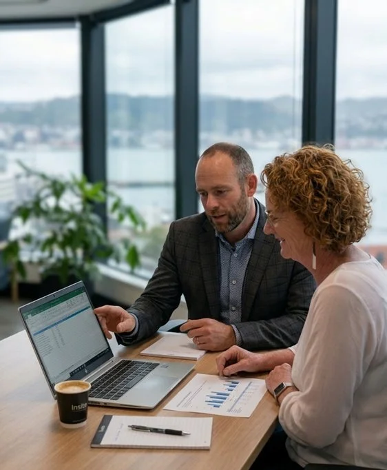 A man and woman are sitting at a table in an office with large windows. They are looking at a laptop with charts and spreadsheets, discussing work. There are documents, a pen, a coffee cup, and a potted plant on the table.
