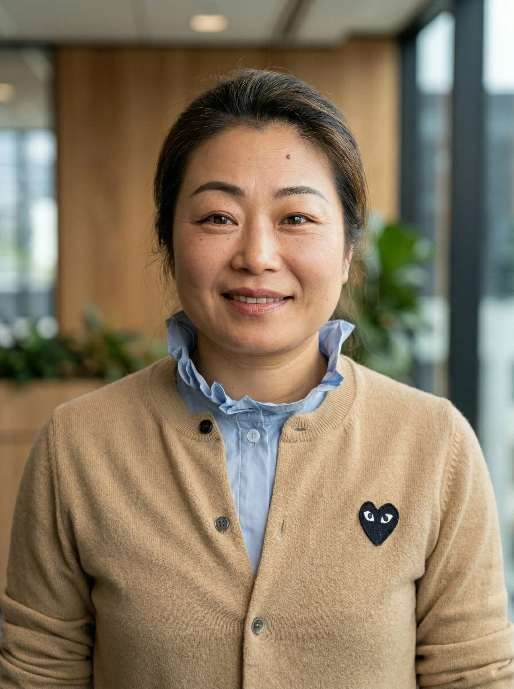 A smiling woman with dark hair pulled back, wearing a beige cardigan with a black heart-shaped logo on the chest, over a light blue shirt with a ruffled collar, standing indoors near large windows with a blurred background of plants.