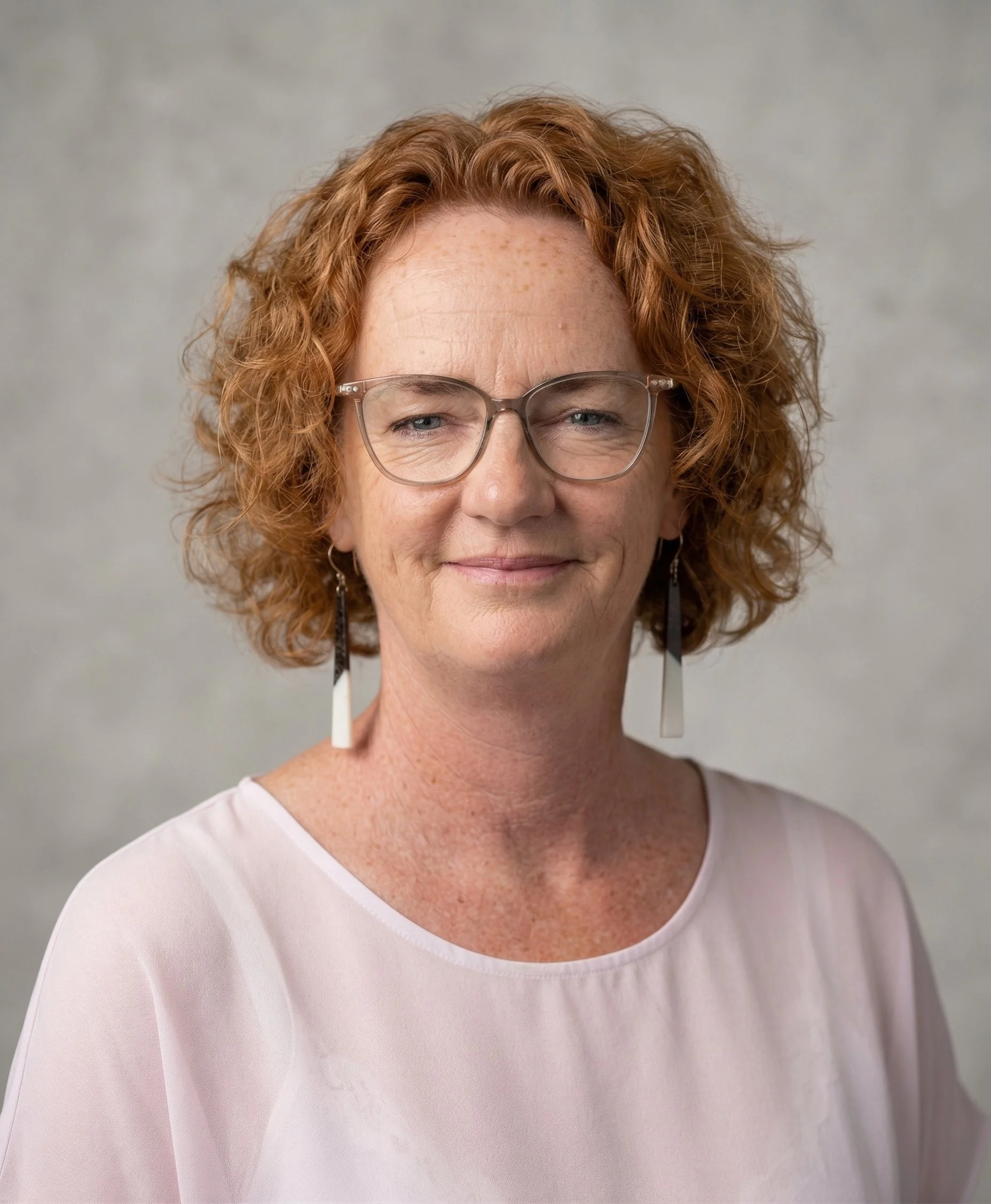 A woman with red curly hair, wearing glasses and long earrings, smiling softly in front of a plain, light background.