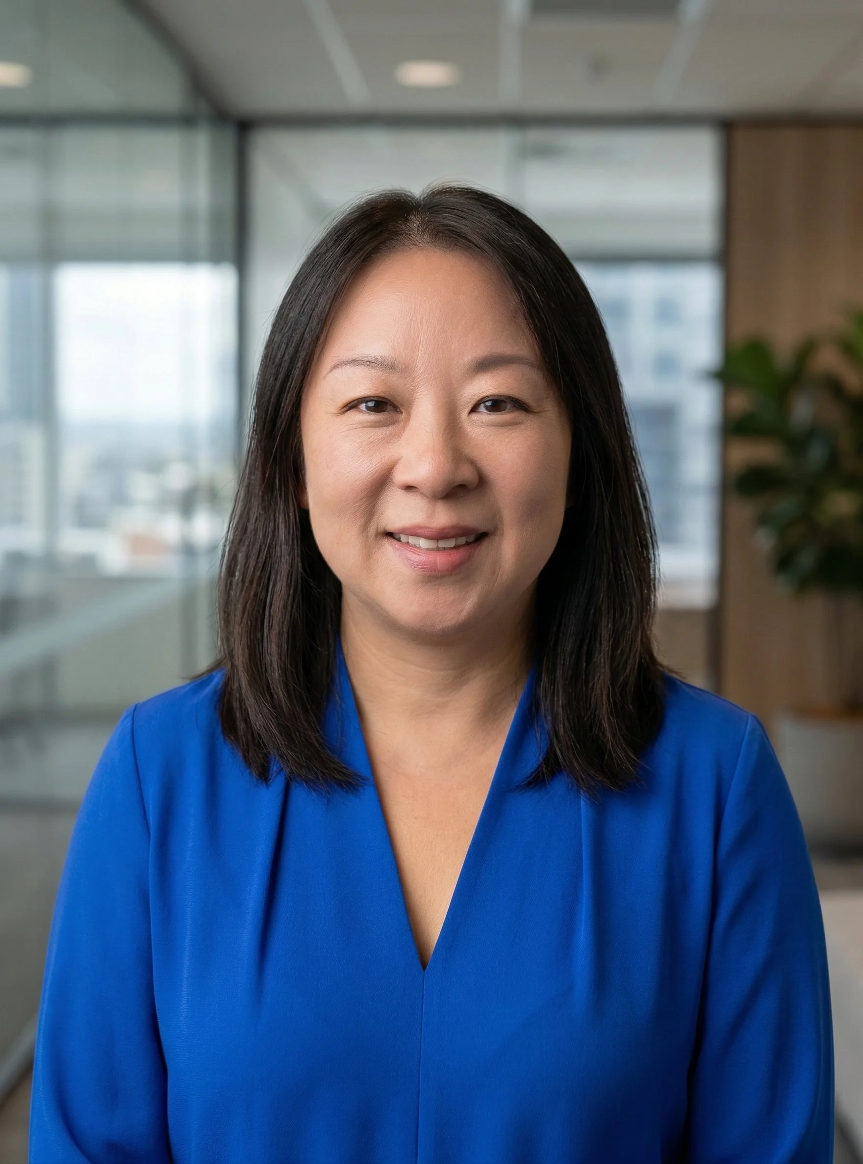 Professional woman with dark hair wearing a blue blouse, smiling, in an office setting with large windows and plants.