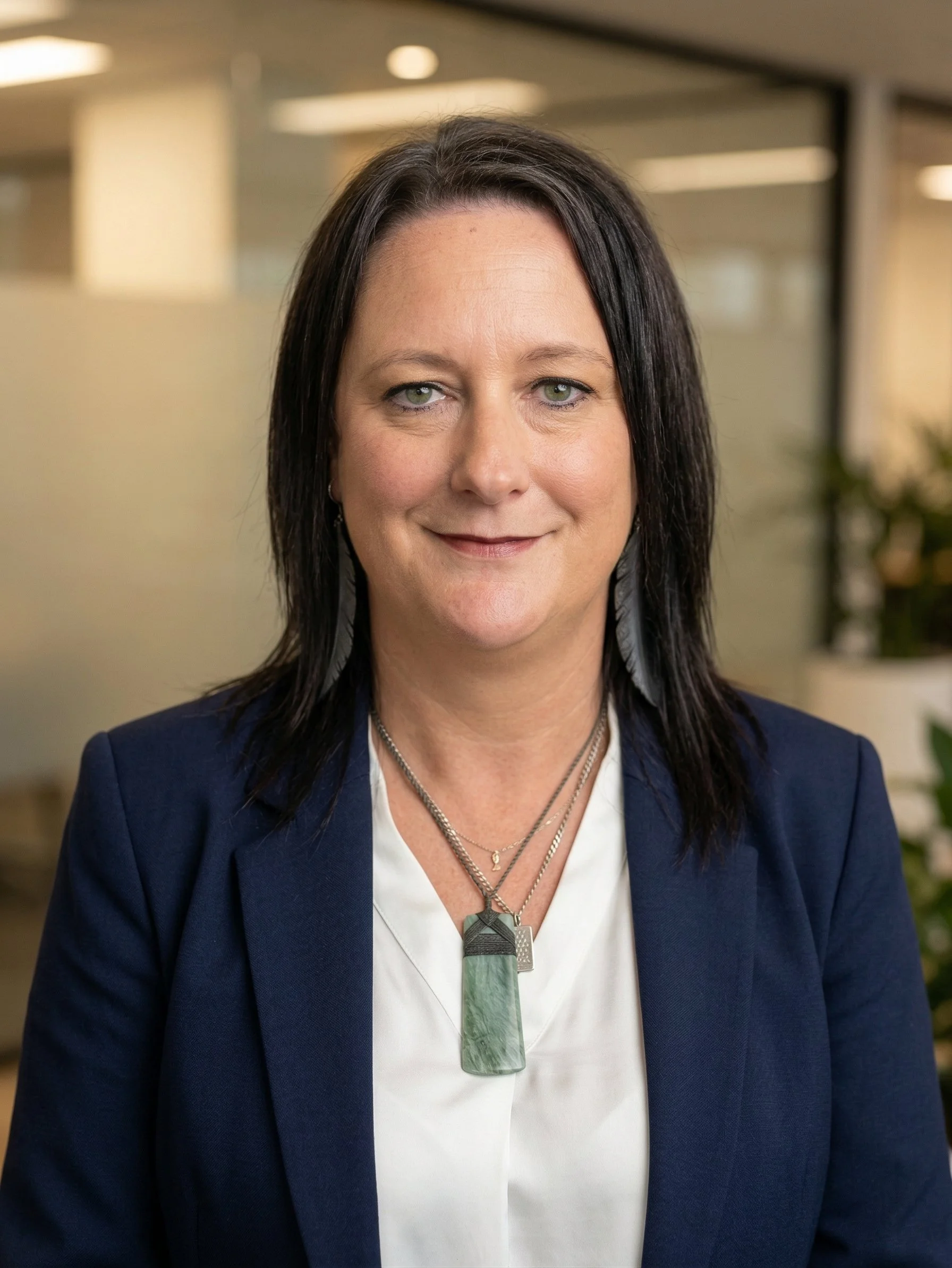 A woman with dark brown hair, wearing a navy blazer and a white blouse, standing in an office environment with plants and lighting fixtures in the background.