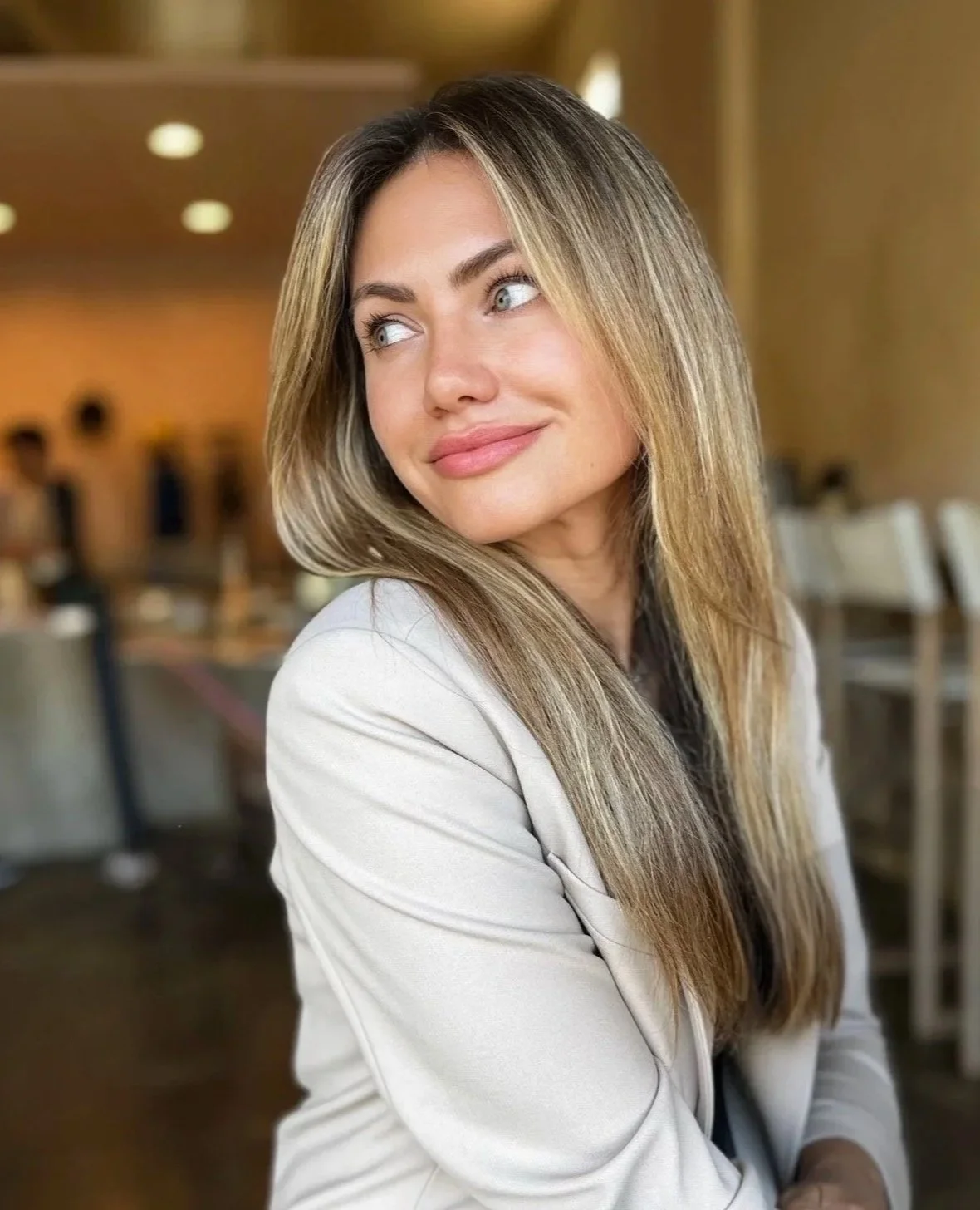 A woman with long blonde hair and blue eyes, wearing a light-colored blazer, smiling and looking to her left, in a warmly lit indoor setting.