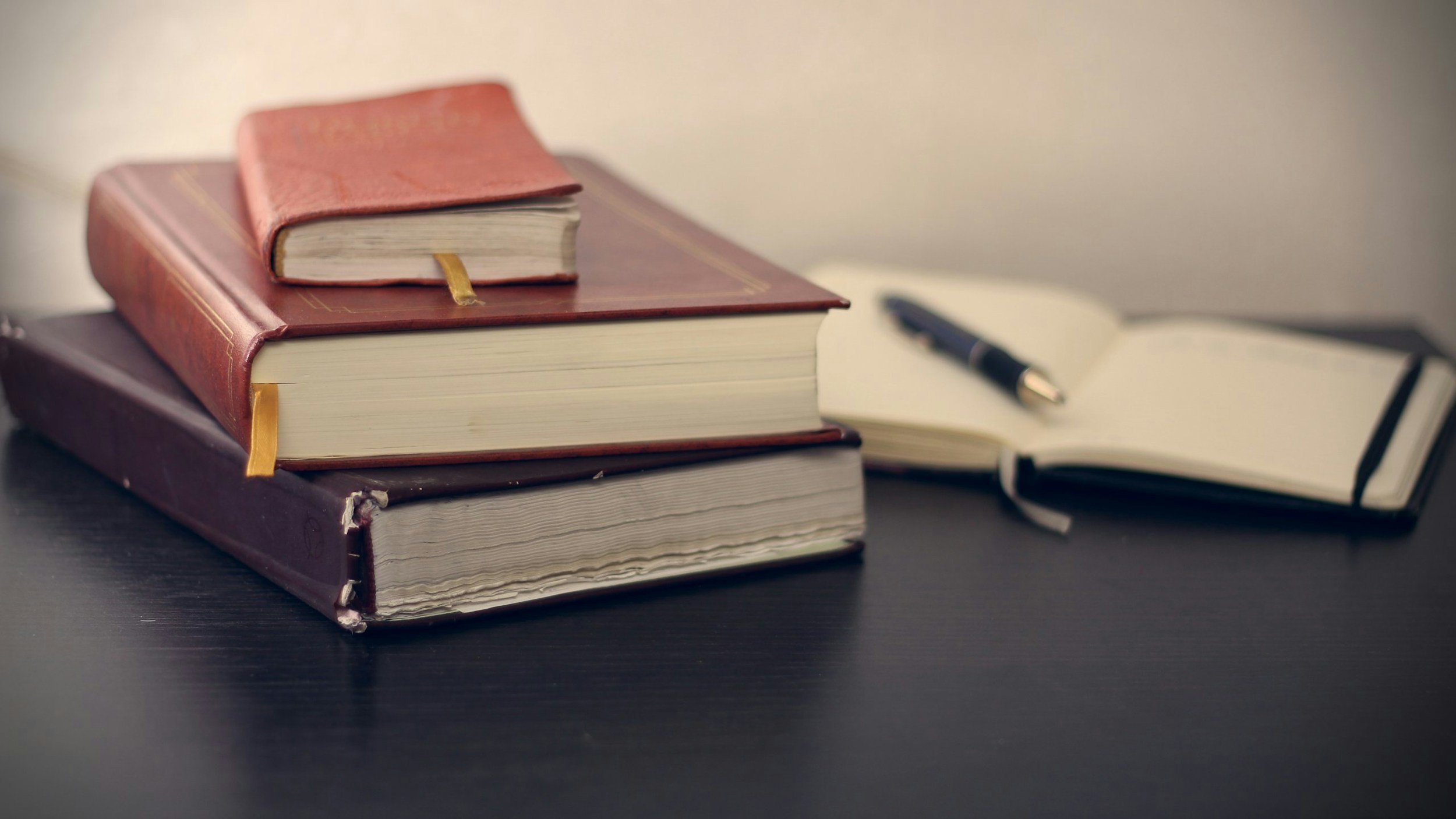 A stack of books with two notebooks, one open and one closed, on a dark surface with a pen nearby.