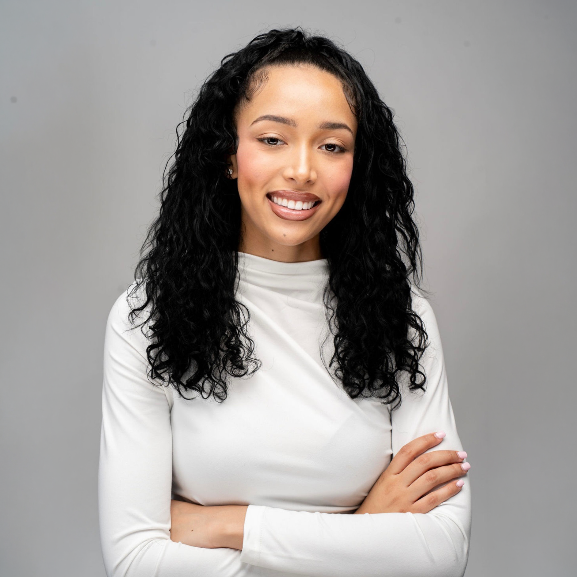 A woman with long curly black hair, wearing a white long-sleeve top, smiling slightly, standing against a gray background with her arms crossed.