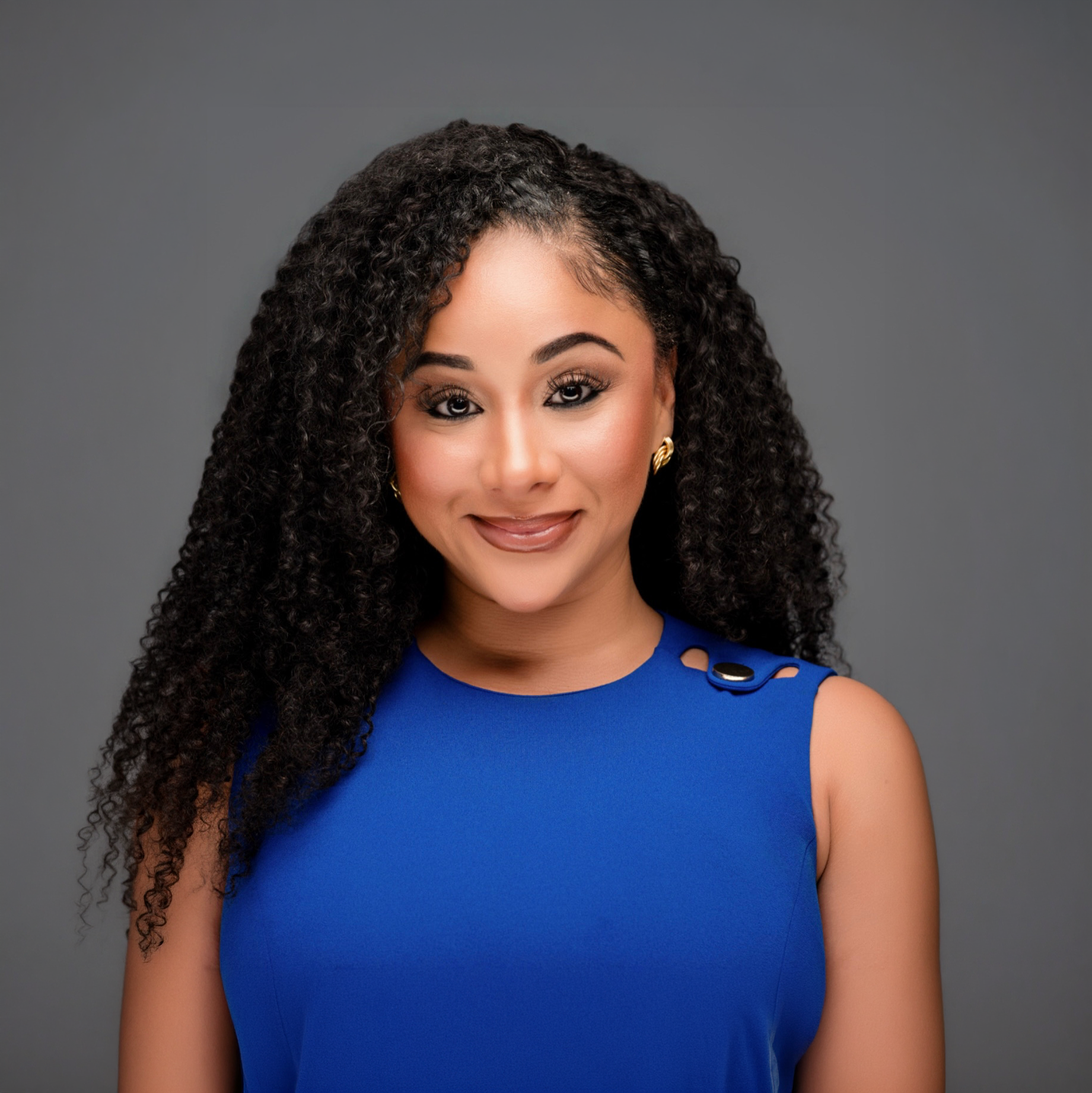 Close-up portrait of a woman with long curly black hair, wearing gold earrings, a bright blue sleeveless dress with button details on the shoulder, and smiling against a plain gray background.