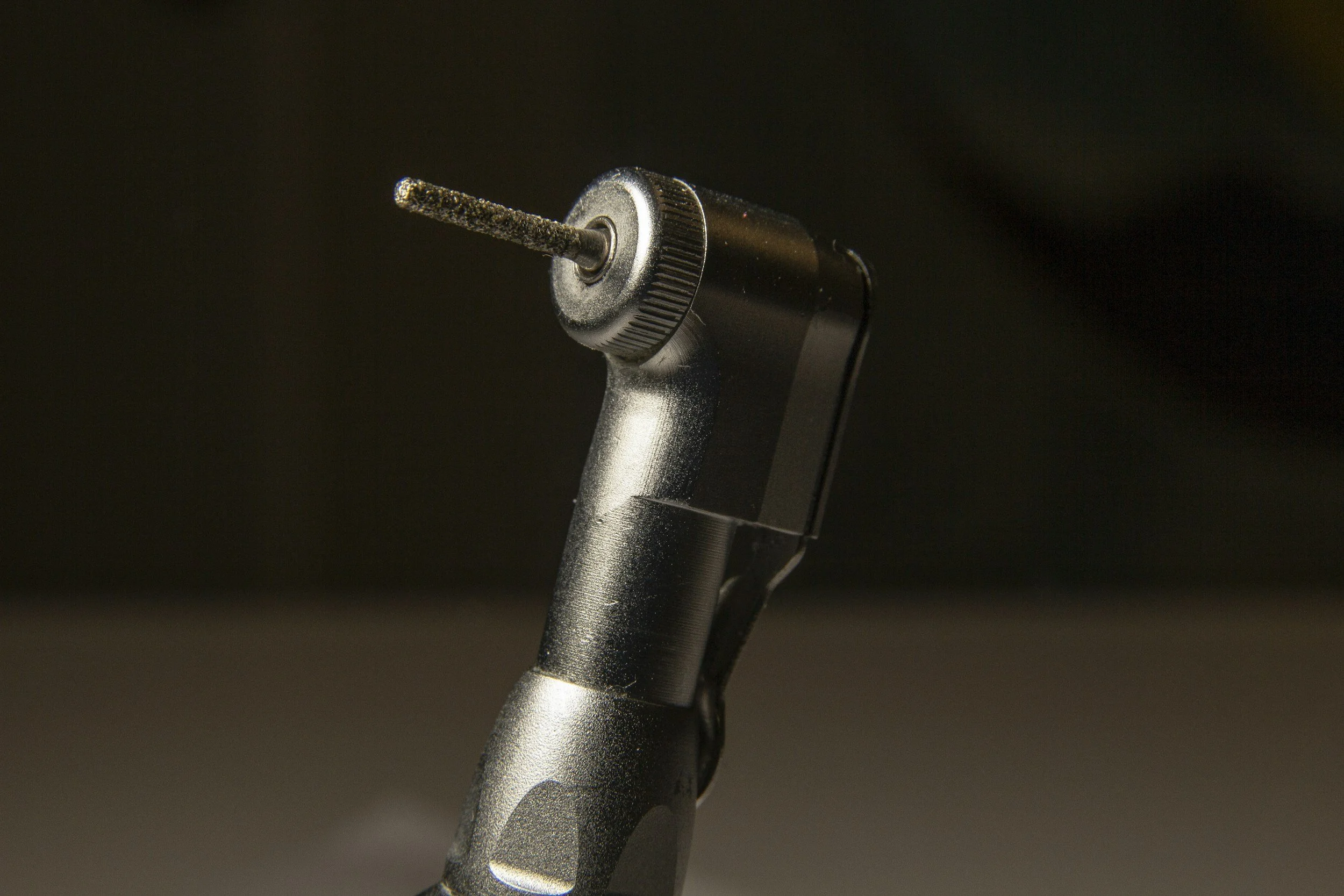Close-up of a black dental handpiece with a bur attached, set against a dark background.