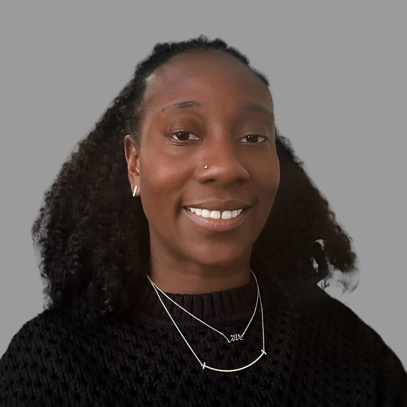 A woman with dark skin and curly hair smiling, wearing layered necklaces and earrings, dressed in a black textured top, against a plain gray background.