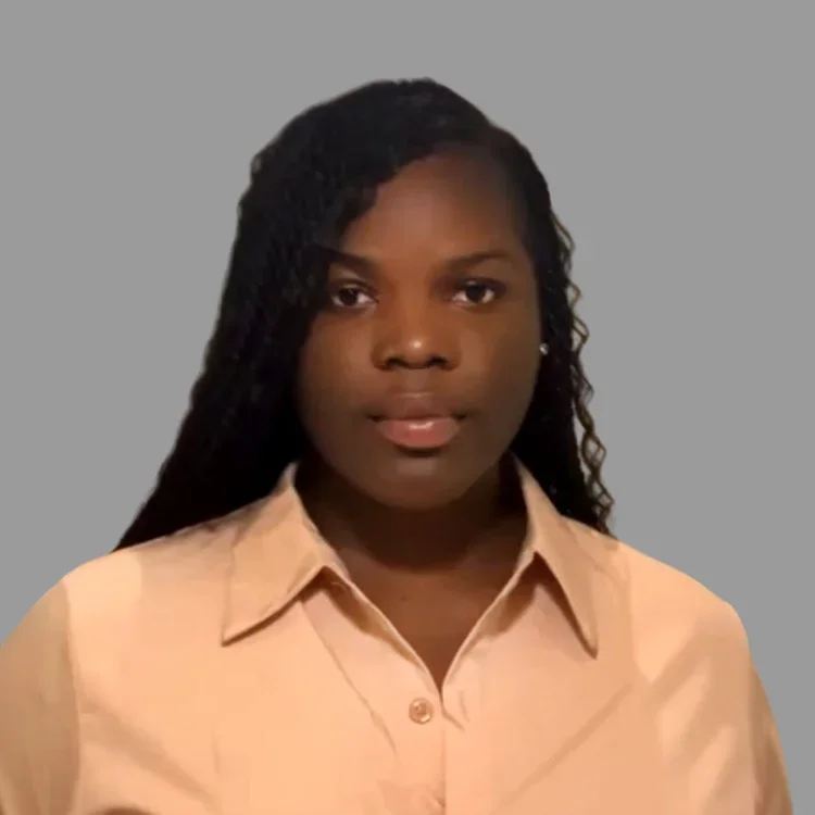 A woman with dark hair styled in twists, wearing a tan button-up shirt, standing against a plain gray background.