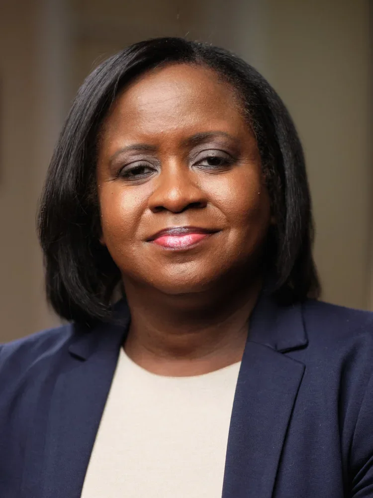 A portrait of an African American woman with shoulder-length black hair, wearing a navy blazer and a beige top, smiling softly at the camera.