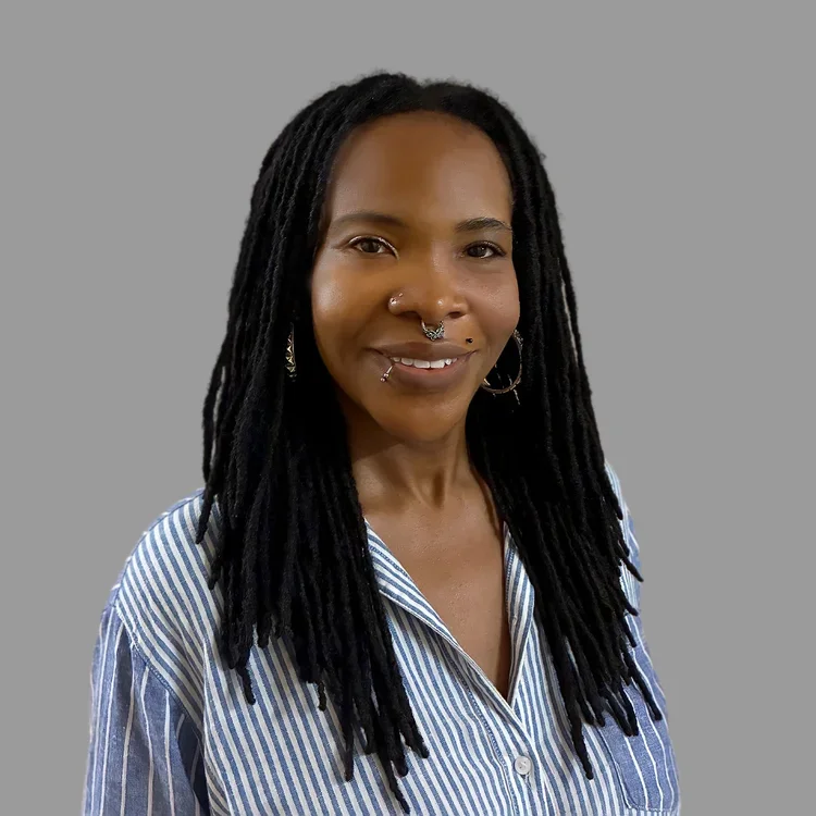 A woman with long black dreadlocks and multiple facial piercings smiling at the camera, wearing a blue and white striped button-up shirt, against a plain gray background.