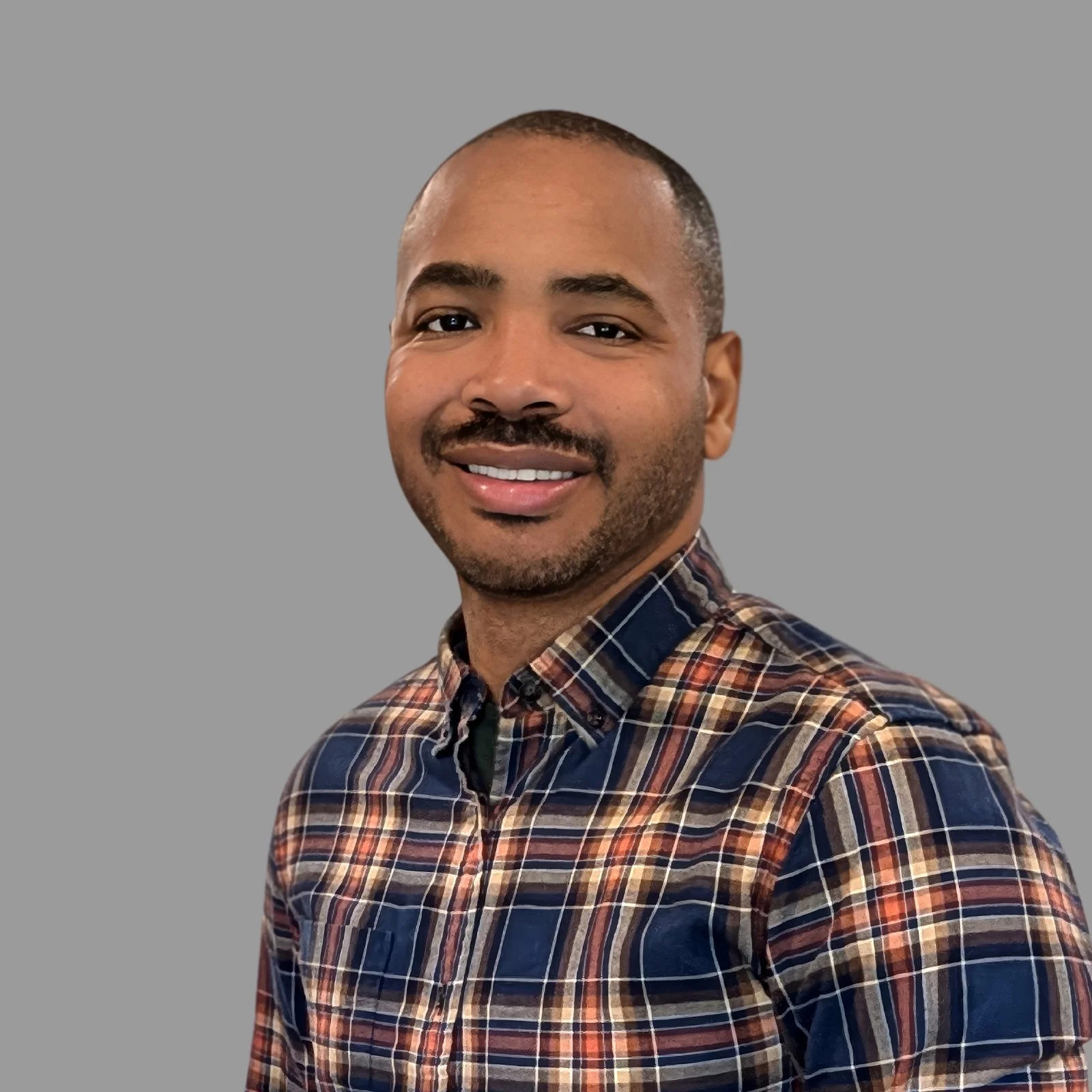 A smiling man with short hair and a beard, wearing a plaid shirt, standing in front of a plain gray background.