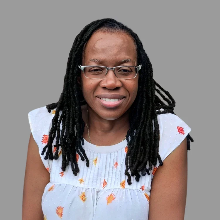 A  black woman with glasses and dreadlocks smiling at the camera against a plain gray background.