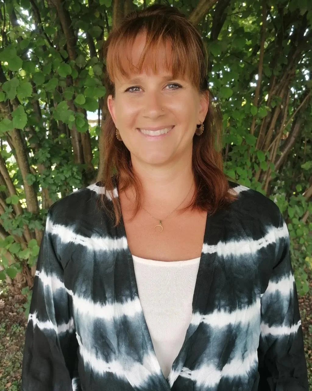 A woman with reddish-brown hair smiling outdoors in front of green leafy bushes, wearing a black and white tie-dye shirt, earrings, and a necklace.