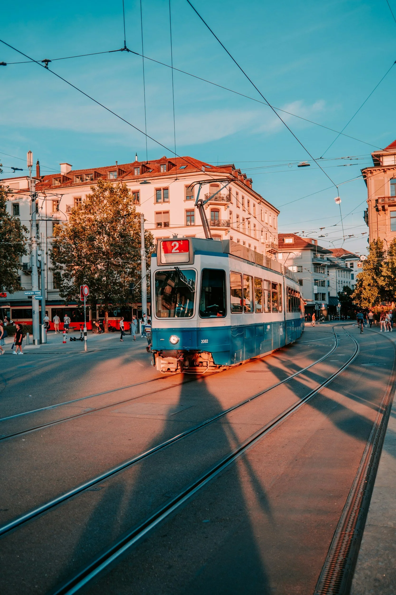 A city street scene in Zurich with a blue and white tram on tracks, overhead wires, pedestrians, and multi-story buildings with red-tiled roofs and trees.