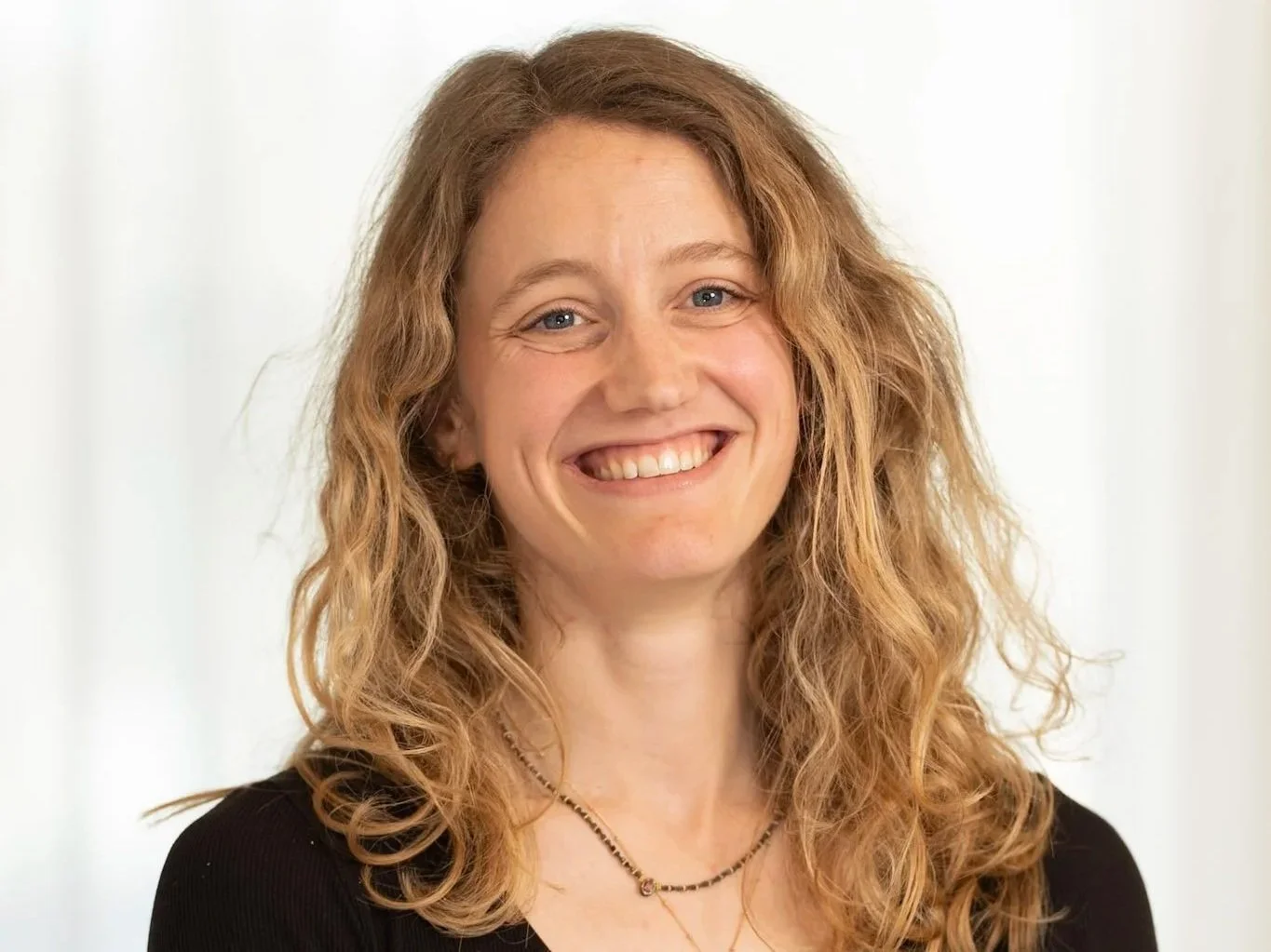 A woman with long, wavy, blonde hair smiling and wearing a black top and a necklace.