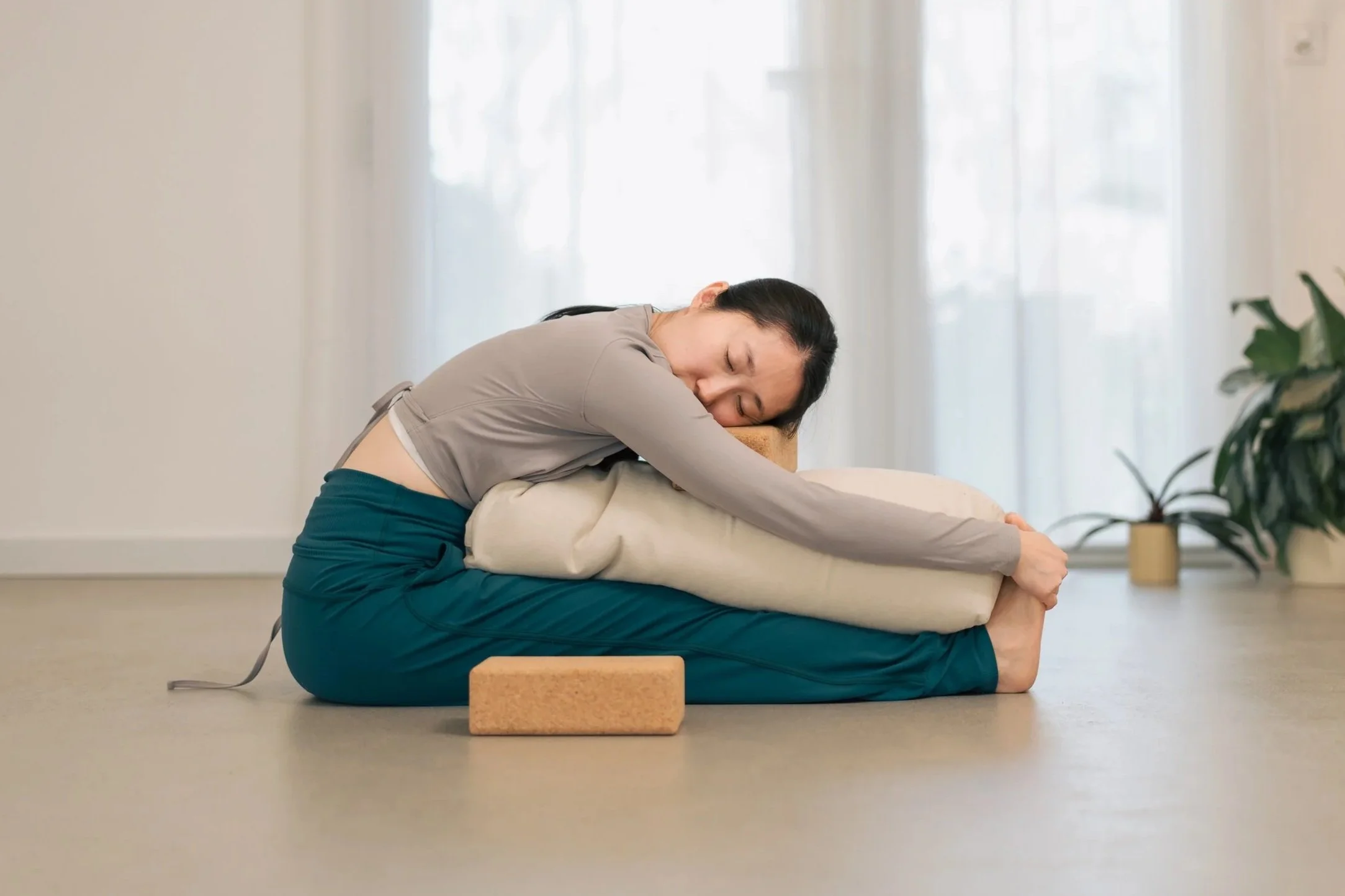 A woman in a yoga class performing a twist on a light-colored wooden floor, wearing a white tank top and light green ribbed leggings.