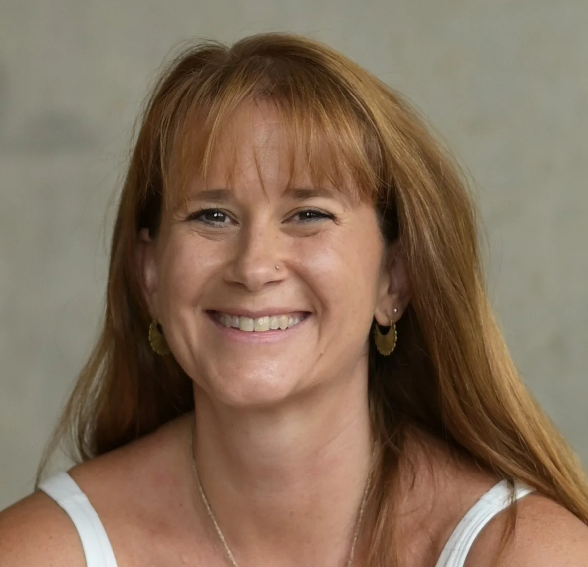 A woman with reddish-brown hair smiling outdoors in front of green leafy bushes, wearing a black and white tie-dye shirt, earrings, and a necklace.