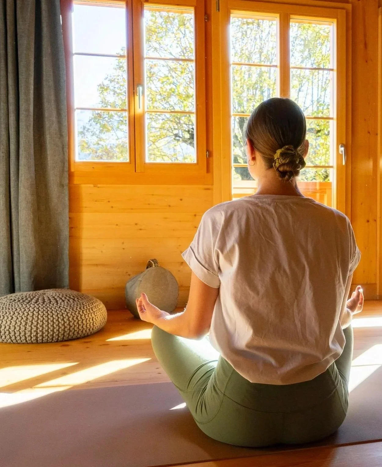 Woman practicing yoga in a hotel with sunlight shining through the windows. In a meditative seated pose.