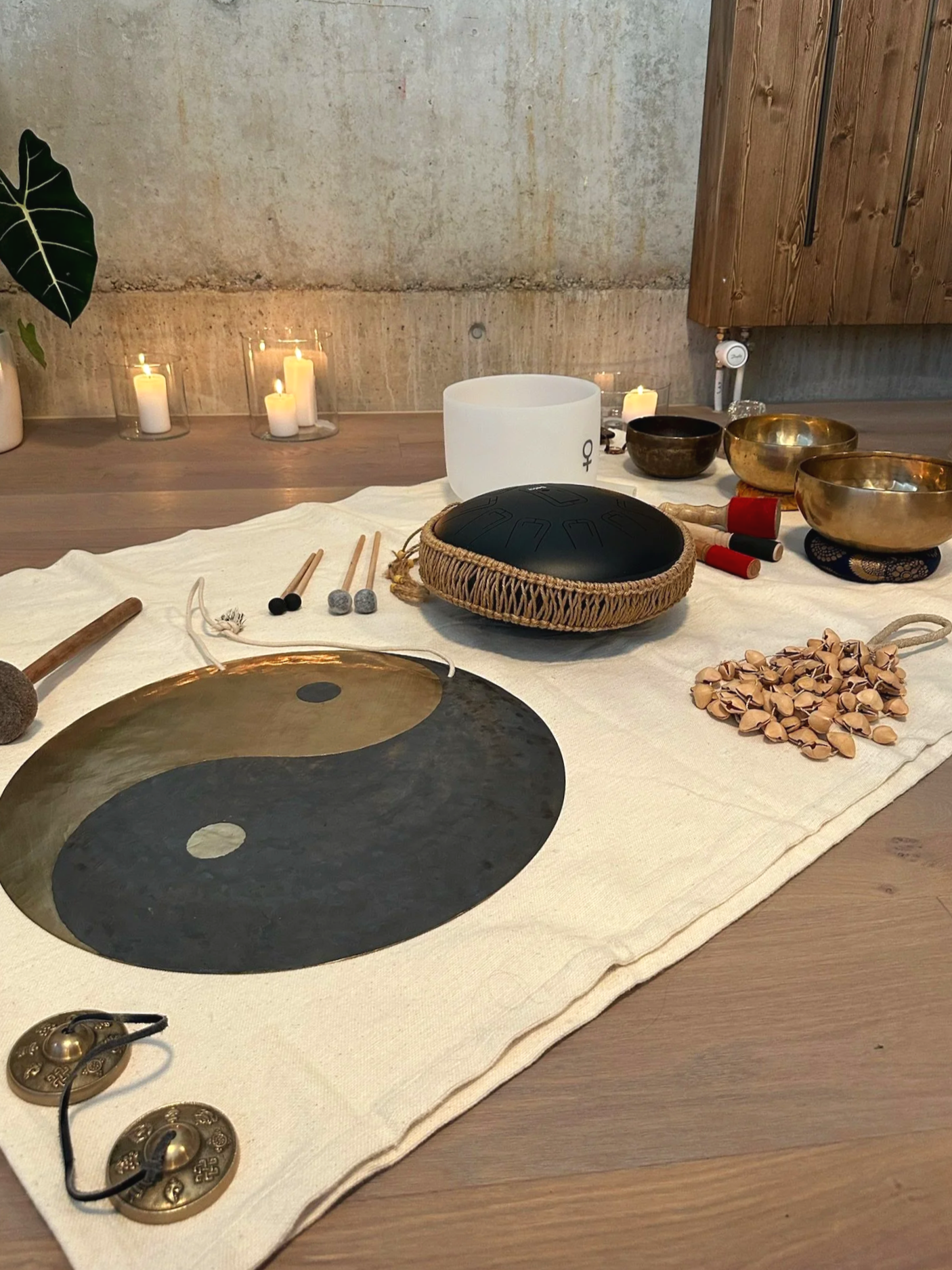 A table with musical instruments including a yin-yang cymbal, singing bowls, mallets, and a white crystal singing bowl, arranged on a white cloth with candles and a plant in the background.