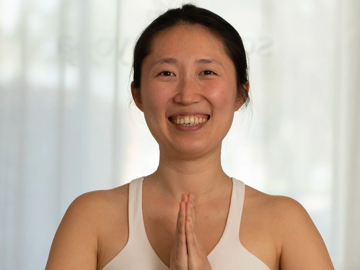 Smiling woman practicing yoga or meditation with hands in prayer pose, wearing a white tank top, in front of a light-colored background.