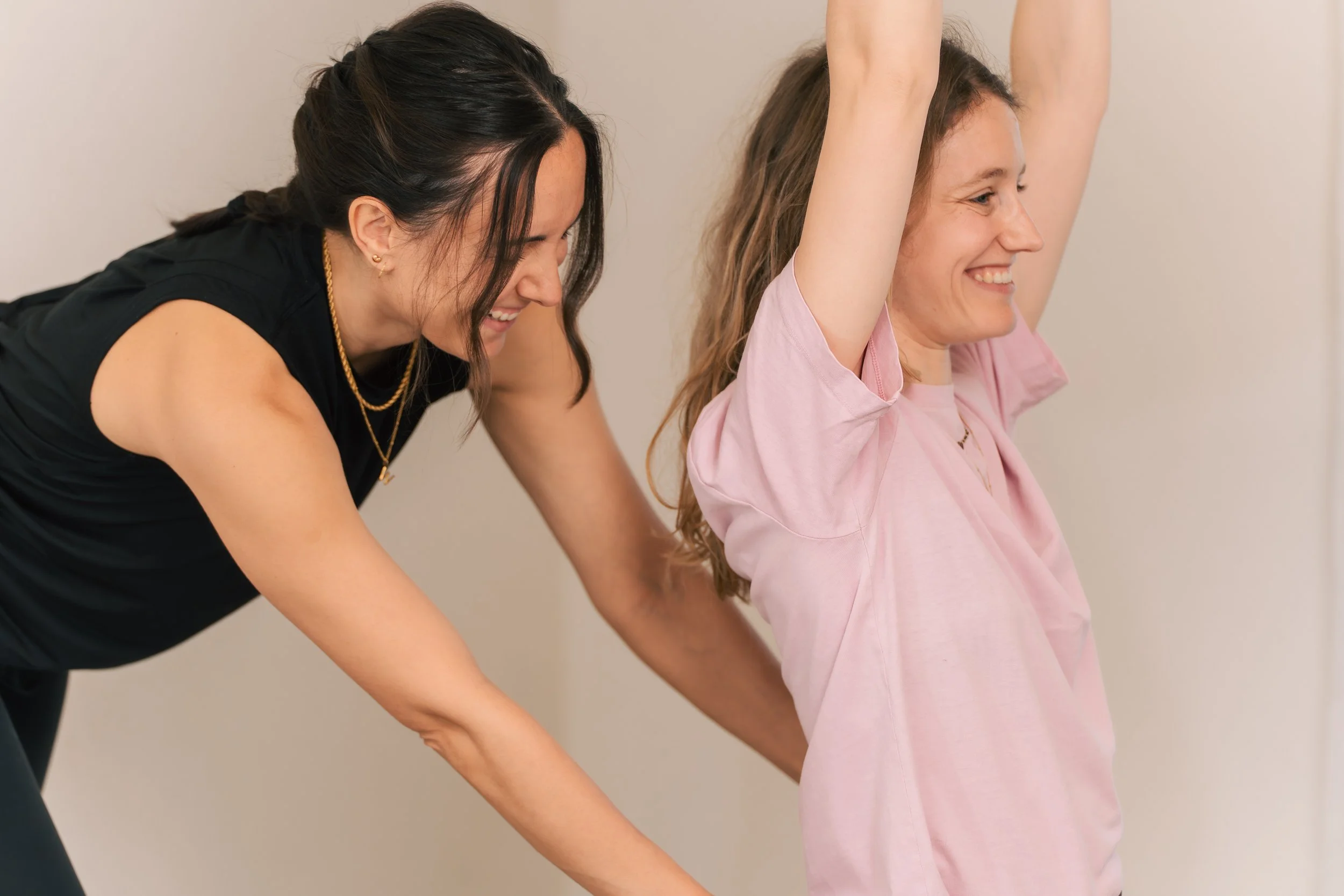 Two women in workout clothes participate in a yoga class, with one woman smiling and raising her arms, and another woman leaning forward to adjust the student.