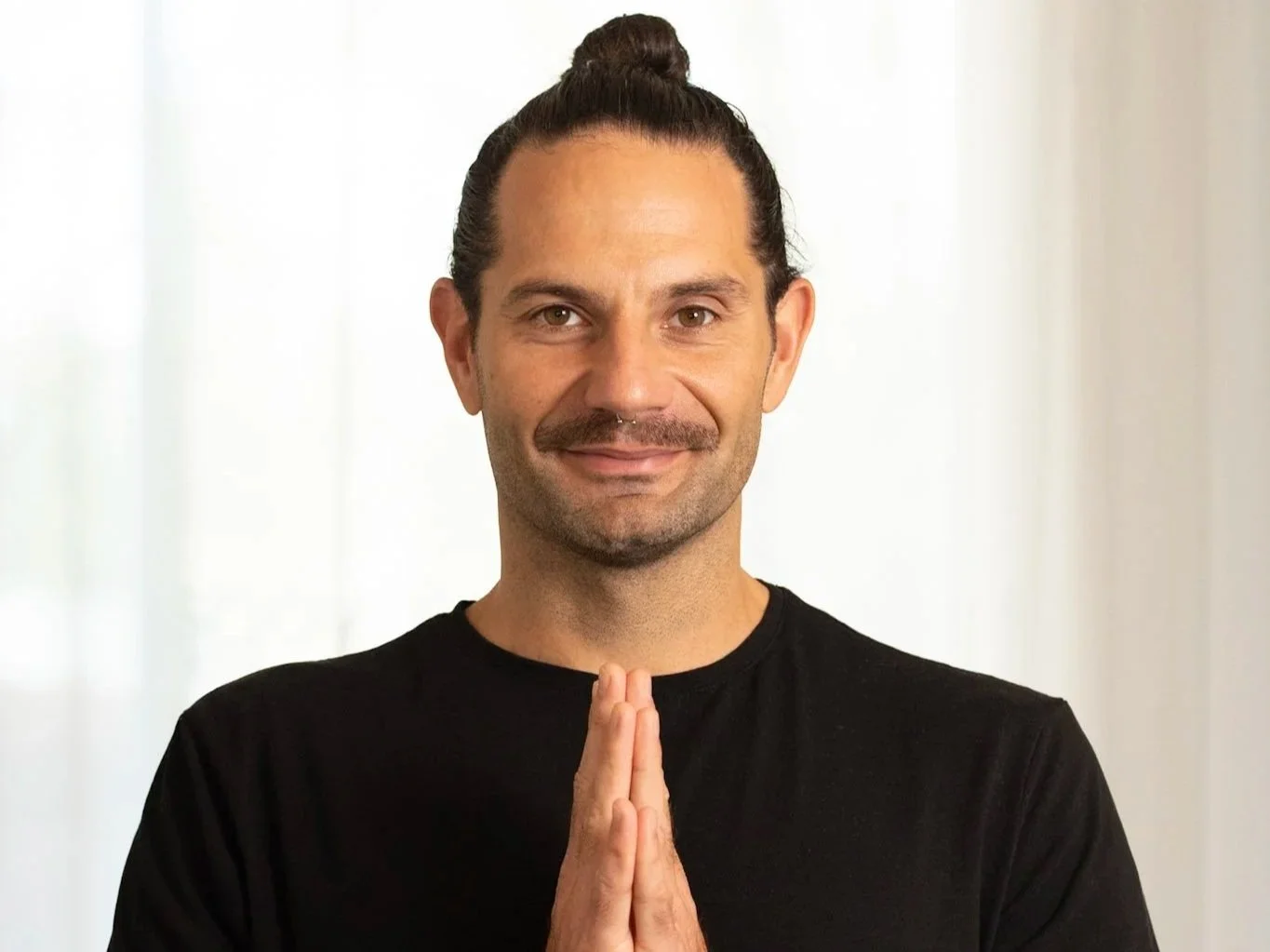 A man with a mustache and a bun hairstyle is smiling at the camera with hands pressed together in a prayer or meditation pose, wearing a black shirt, in a bright, neutral background.
