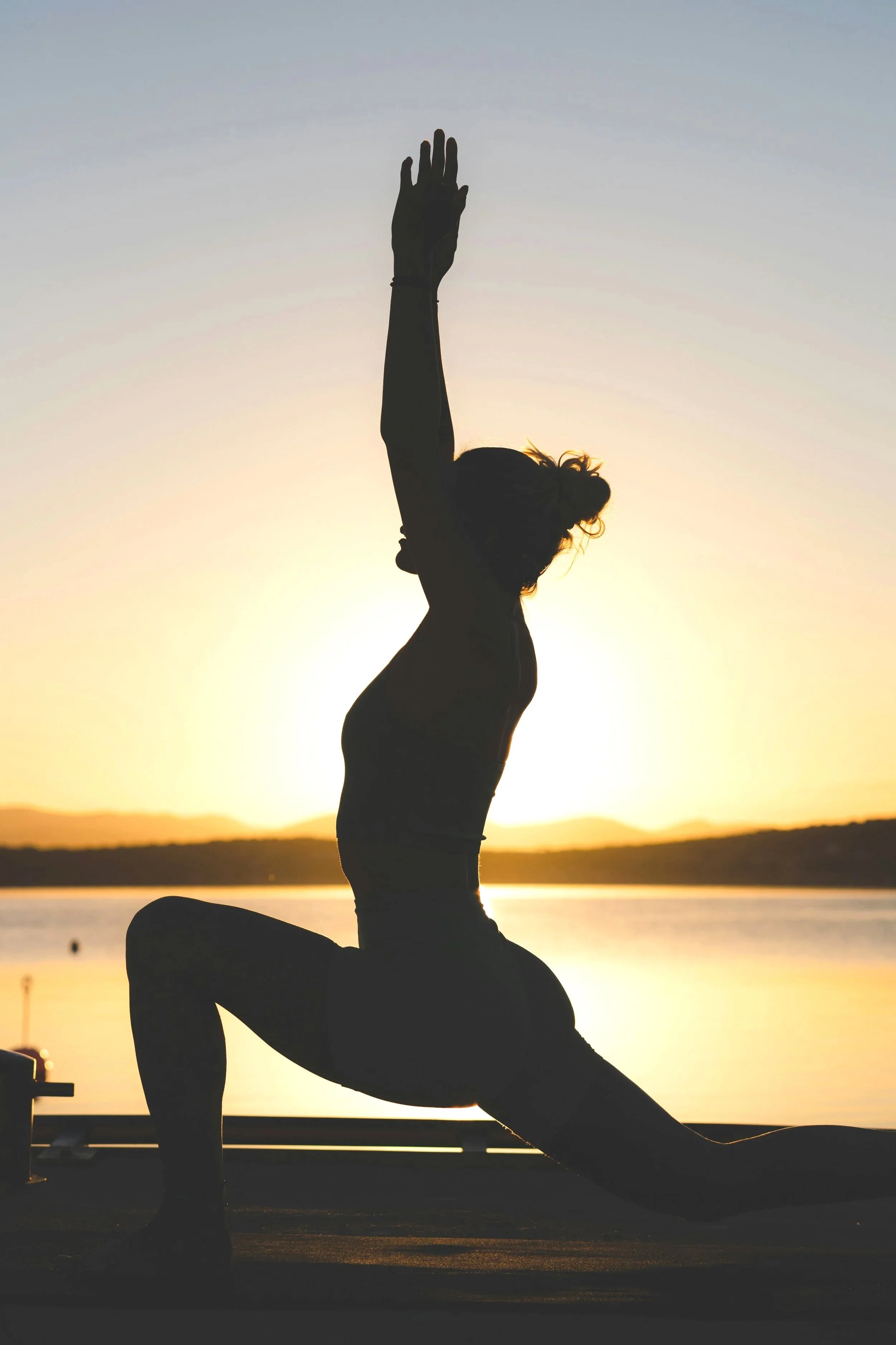 Silhouette of a woman doing yoga in a lunge pose at sunset by the water, with her arms raised overhead.