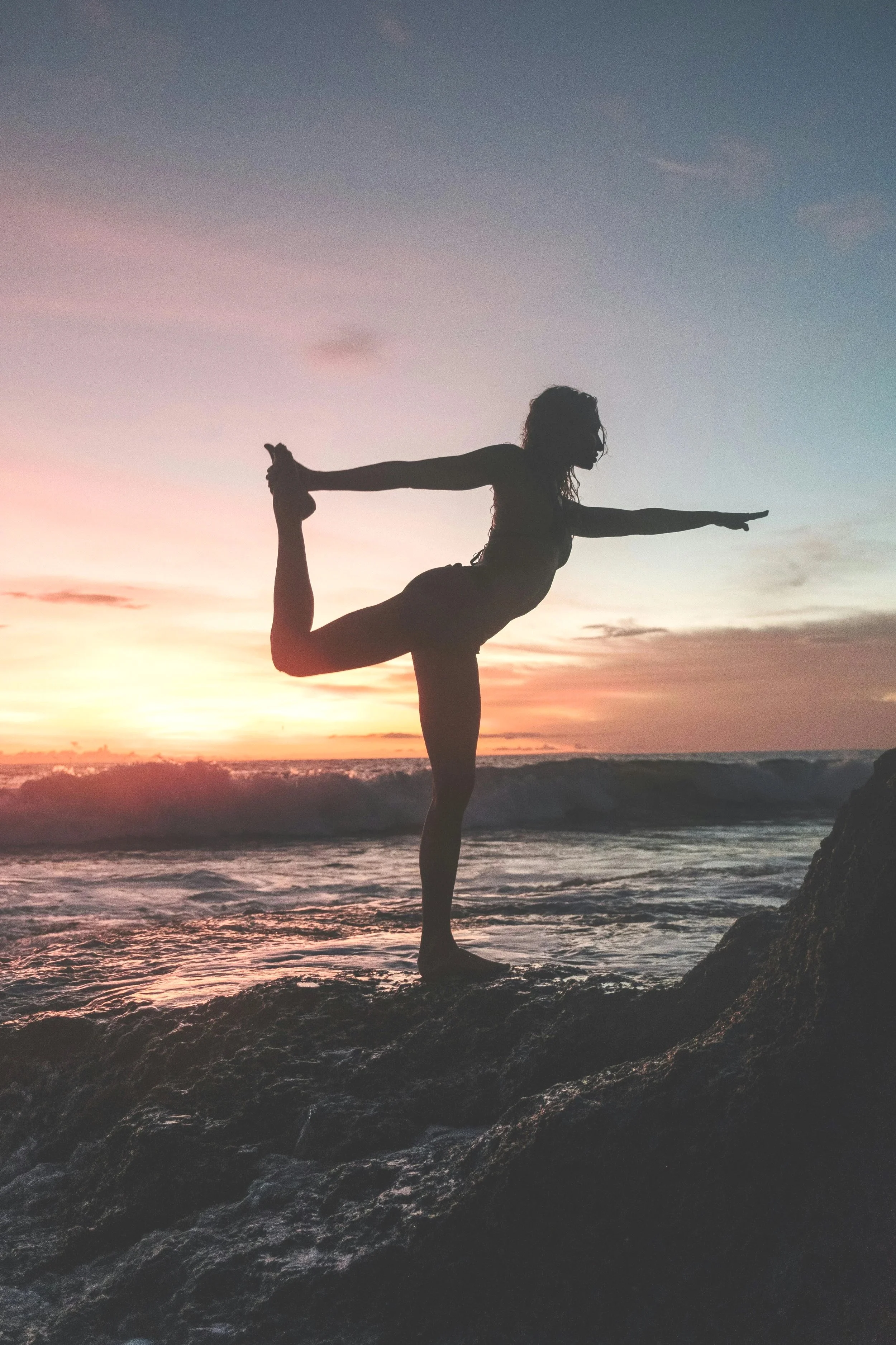 Silhouette of a woman practicing yoga on a rock at the beach during sunset, performing a variation of the dancer pose with one leg bent behind and held by her hand, while extending the other arm forward.