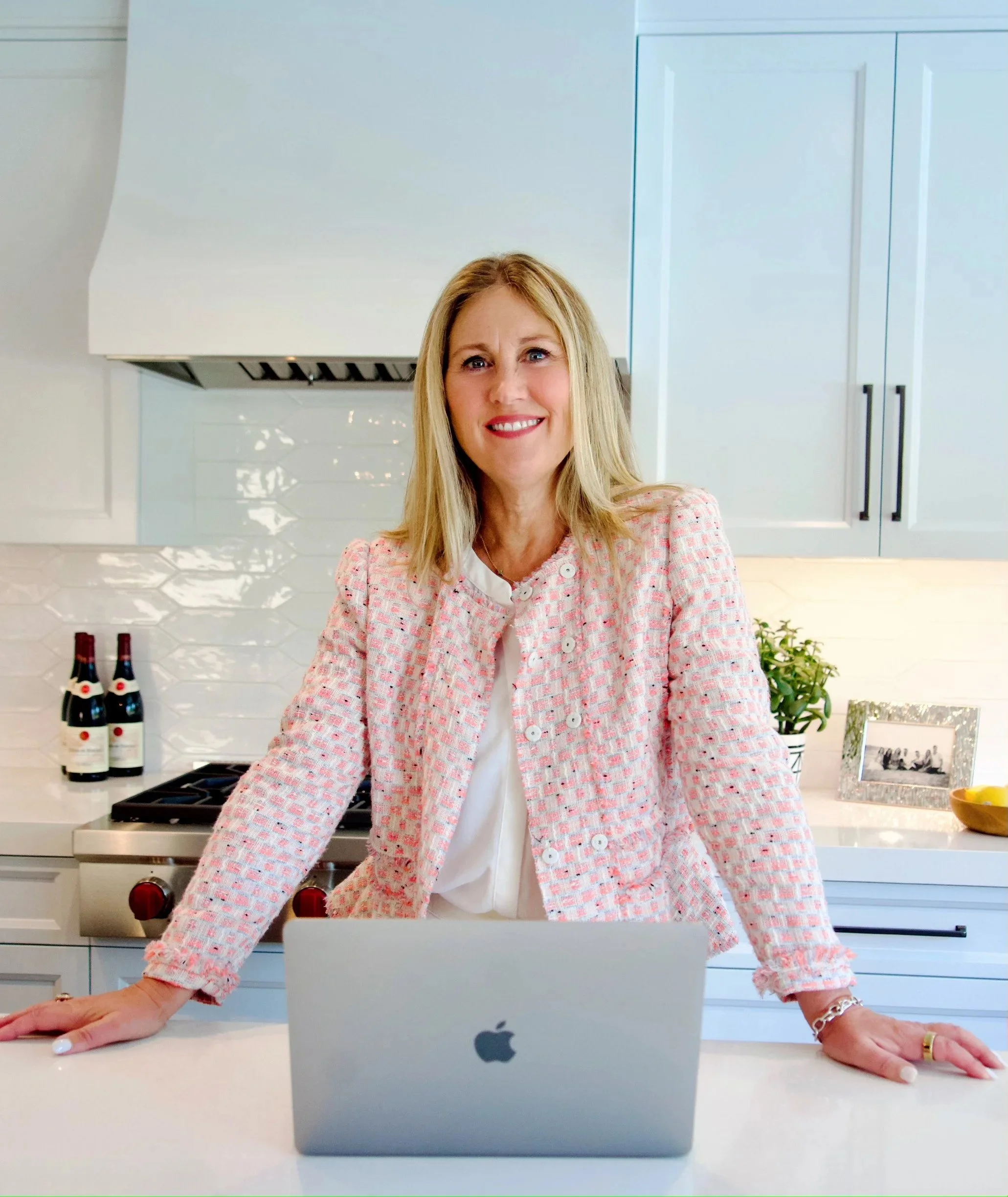 A woman with blonde hair smiling and standing behind a silver MacBook laptop on a kitchen counter.