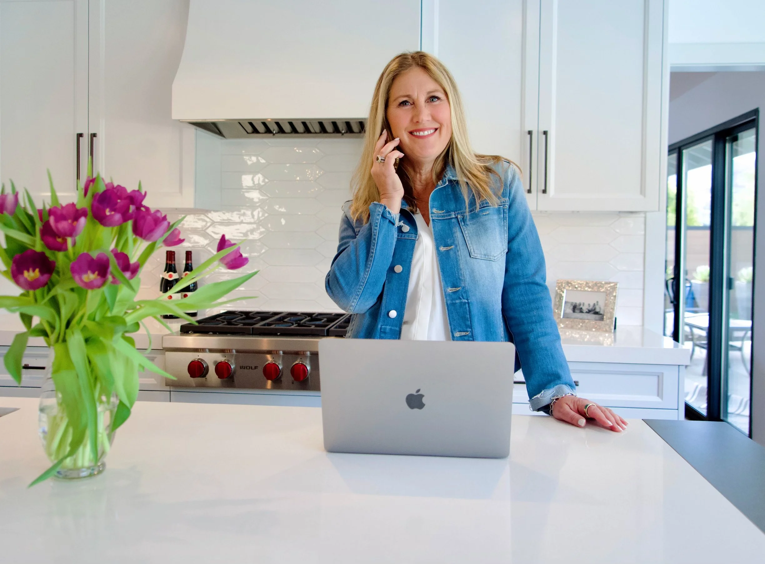 A smiling woman in a denim jacket talking on a phone in a modern kitchen with a laptop, a vase with purple tulips, and a framed photo on the counter.