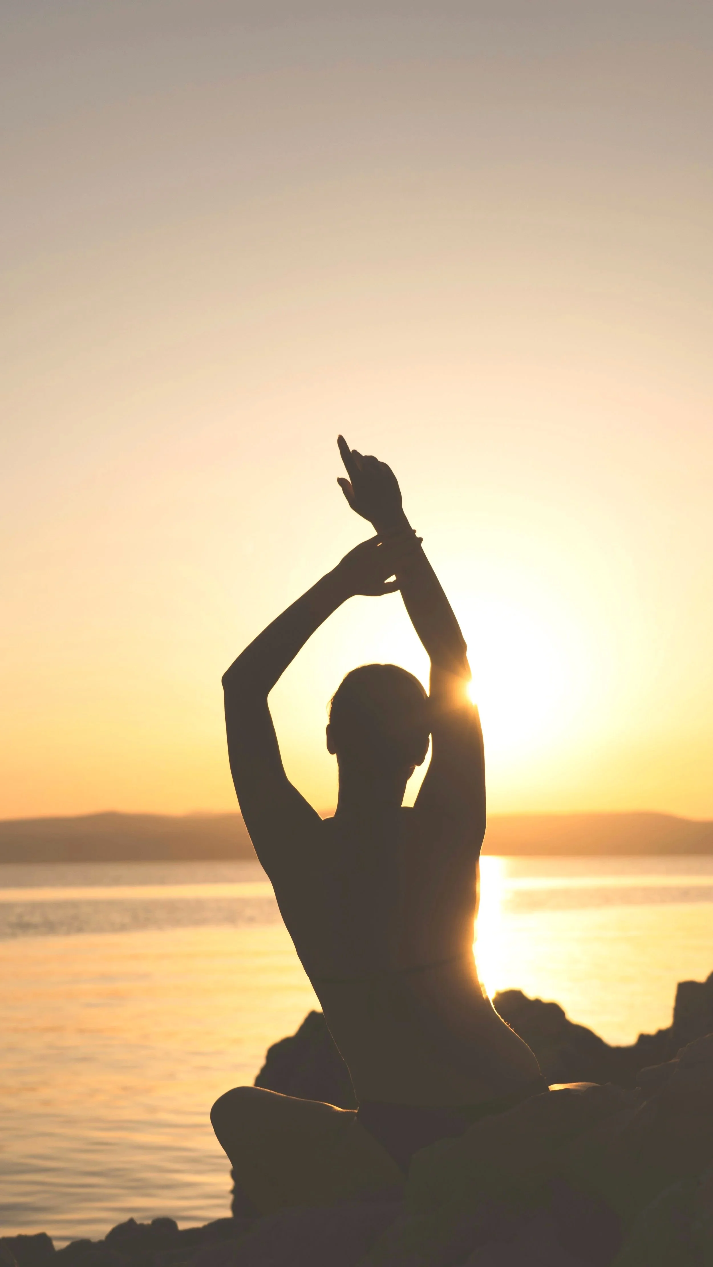 Silhouette of a person meditating on rocks by the water during sunset.