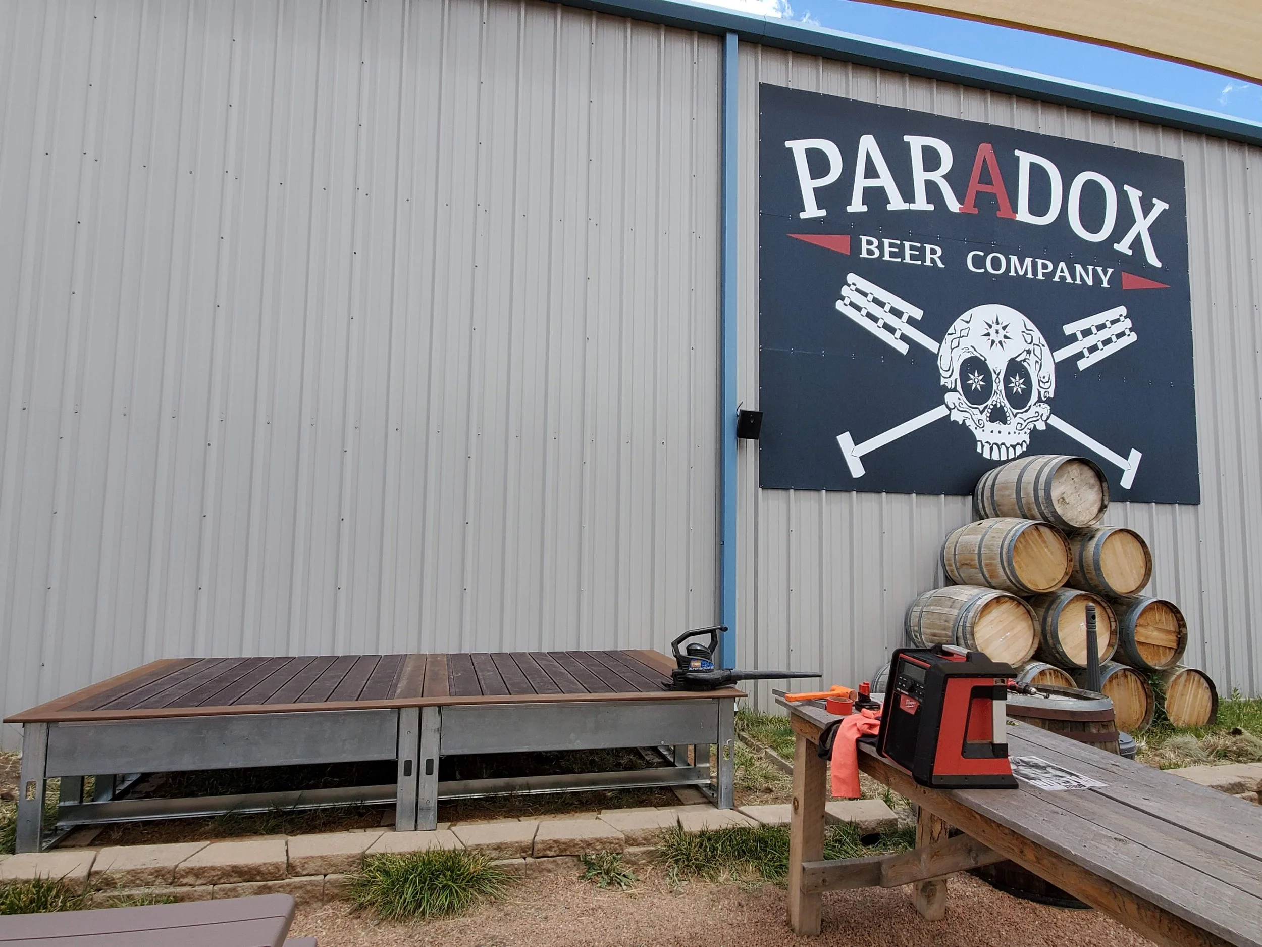 Exterior of a brewery with a large sign for Paradox Beer Company featuring a skull with stars for eyes and crossed beer barrels. There are several beer barrels stacked on the ground, a wooden platform, and various tools and equipment in the foregroun