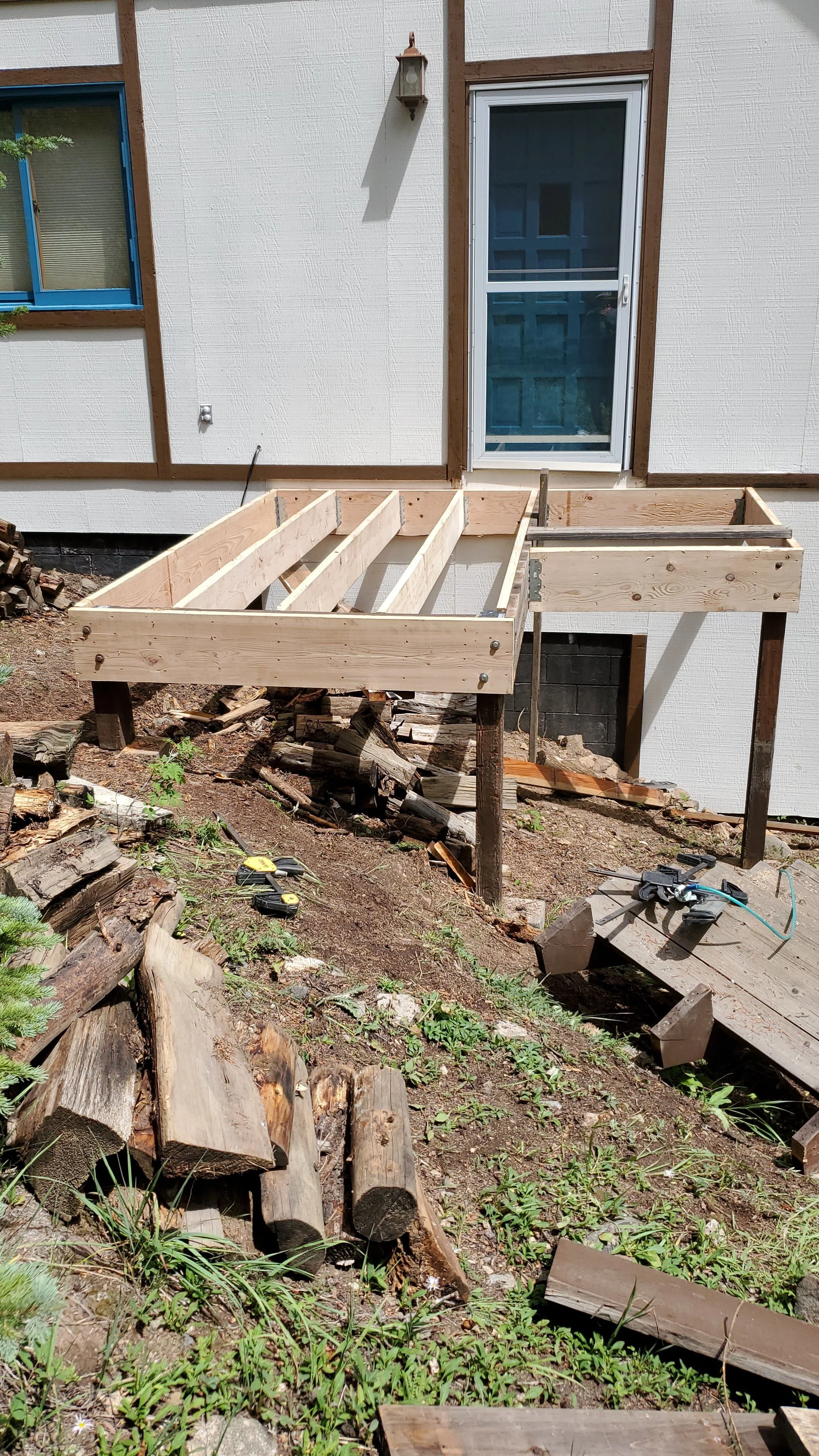 A home exterior with a white wall, a door with a screen, window with blue trim, and an unfinished wooden deck structure being built. Construction tools and wood logs are on the ground.