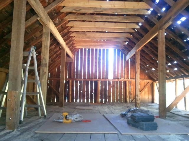 Interior of a wooden barn under construction, showing exposed wooden beams, a ladder, and construction tools on the floor.