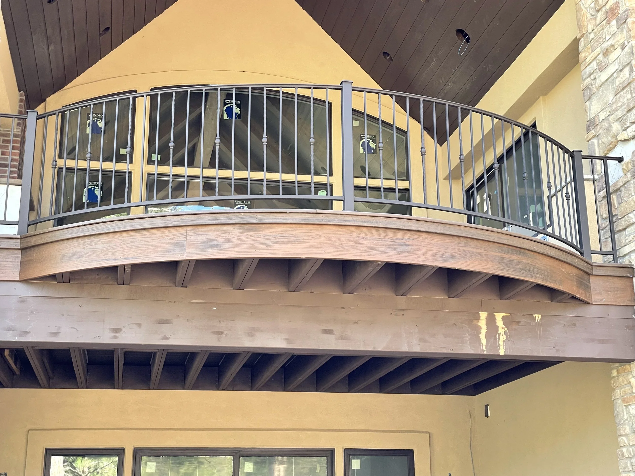 View of a building's second story with a curved black metal balcony railing, large windows with sticker labels, yellow stucco walls, a wooden ceiling, and stone accents.