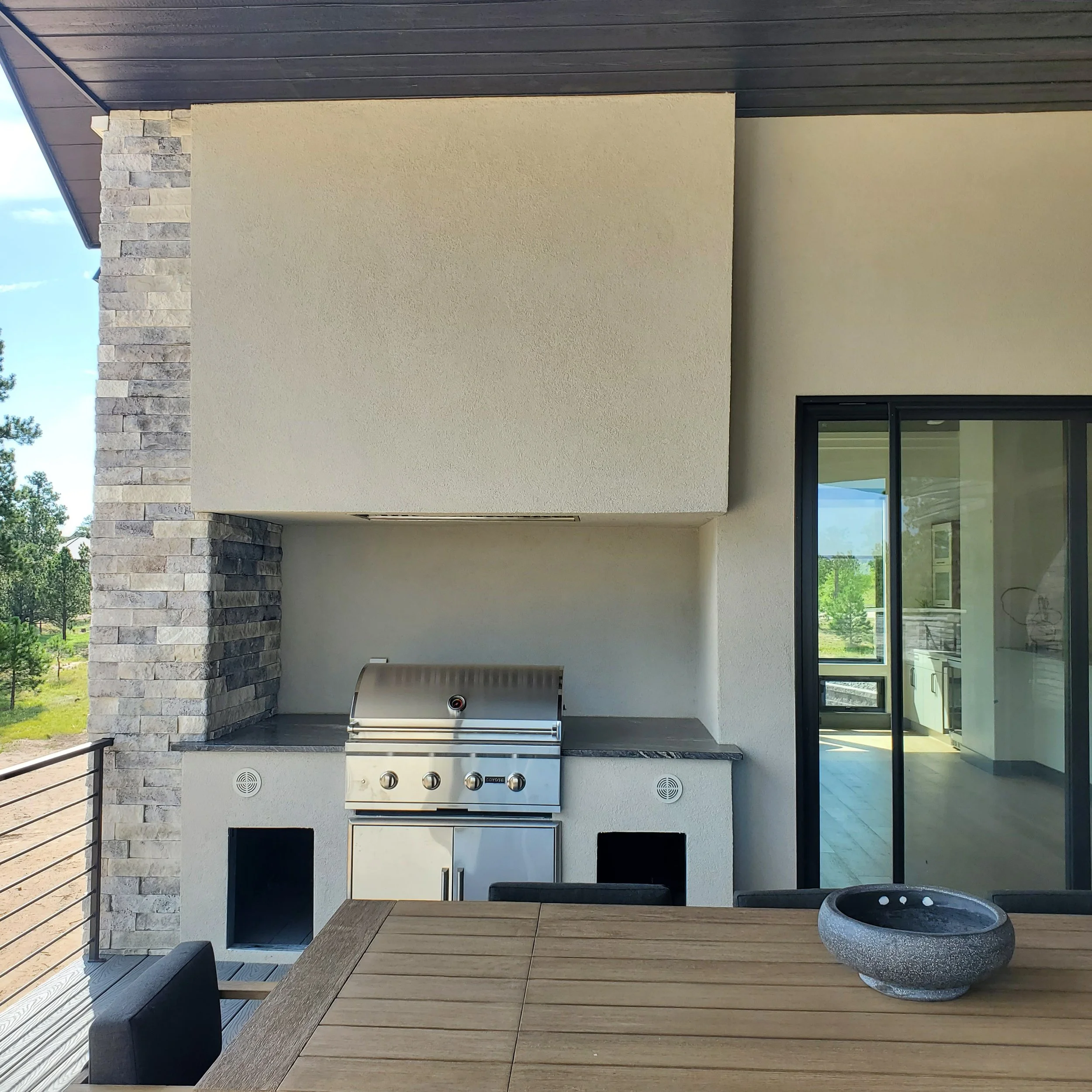 A modern outdoor patio with a built-in stainless steel grill, wooden deck flooring, stone accent wall, and sliding glass doors leading inside. There is a round stone fire pit on the patio.
