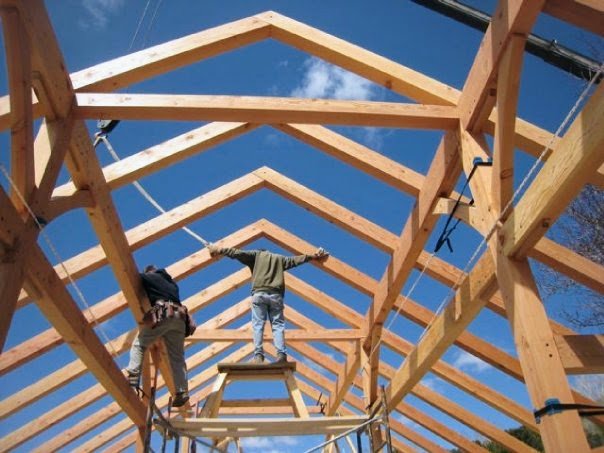 Two construction workers building a timber frame roof with rafters and purlins with a gabled roof against a clear blue sky.