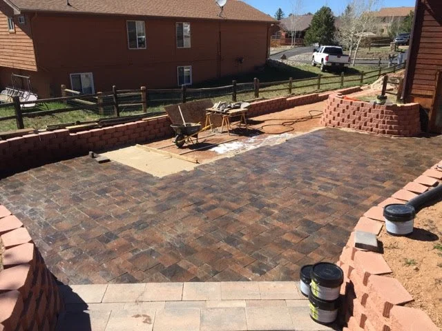 A backyard patio under construction with brick pavers, a wheelbarrow, and stacks of bricks, surrounded by a low brick wall and neighboring houses.