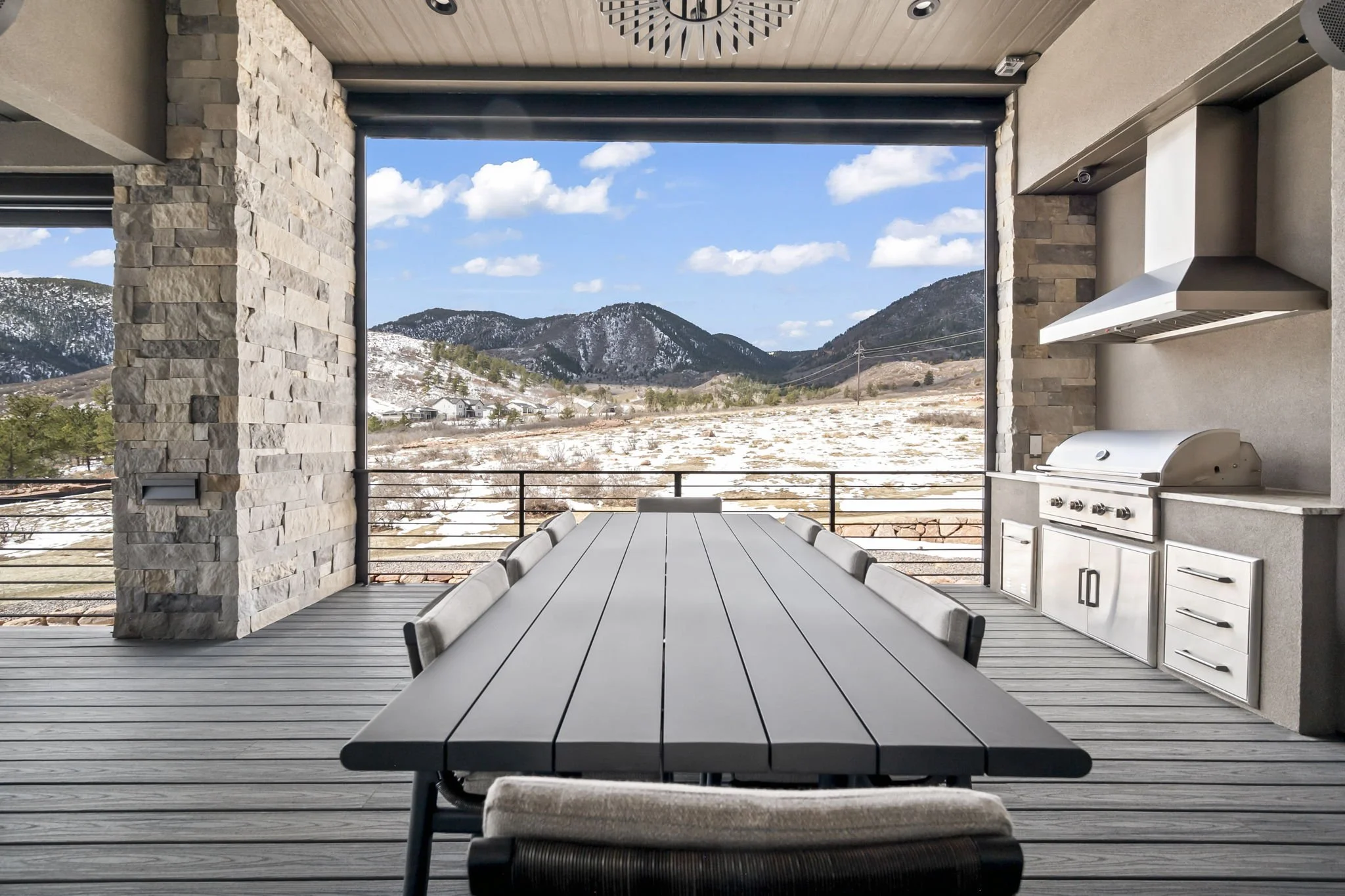 An outdoor Trex deck with a large dining table surrounded by chairs, a built-in barbecue grill, and a view of mountains and a partly cloudy sky in the distance.