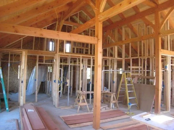 Interior view of a timber frame house under construction with exposed wooden framing, stairs, ladder, and building materials.