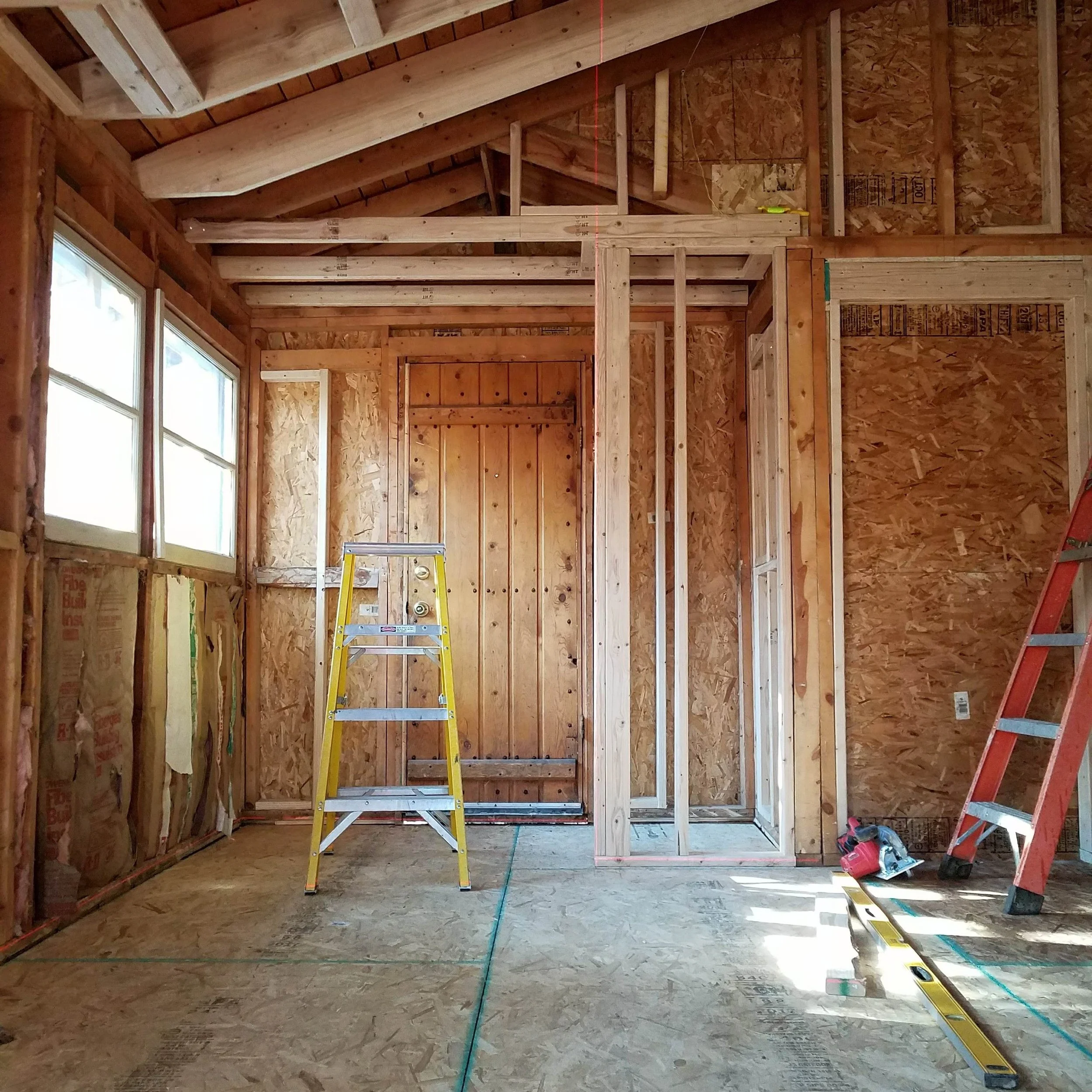 The interior of a house under construction with exposed wooden framing, windows, and tools. A yellow step ladder and a red ladder are visible, along with construction markings on the floor.