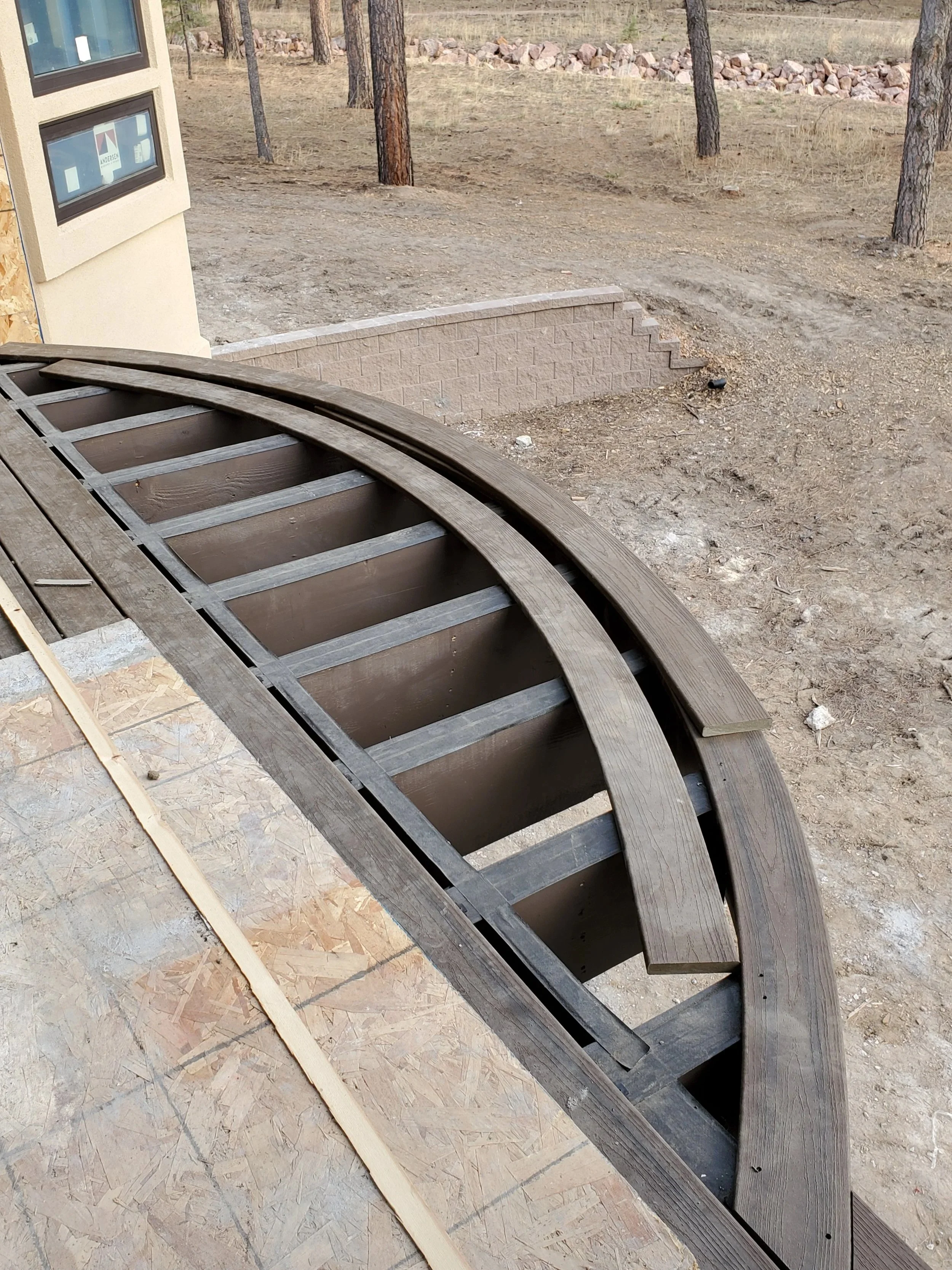 Landscape showing a construction site with a curved wood framed deck on the left, a small brick wall in the center, and a dirt path with trees in the background.