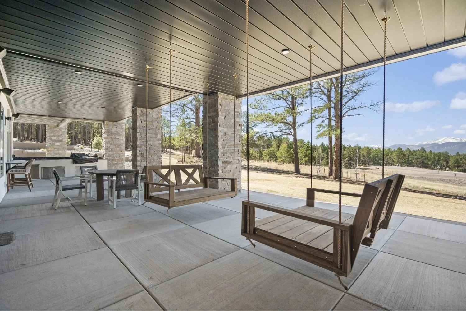 Covered outdoor patio with stone pillar support, wood ceiling, swings, outdoor dining table, chairs, and scenic mountain landscape with trees and blue sky in the background.
