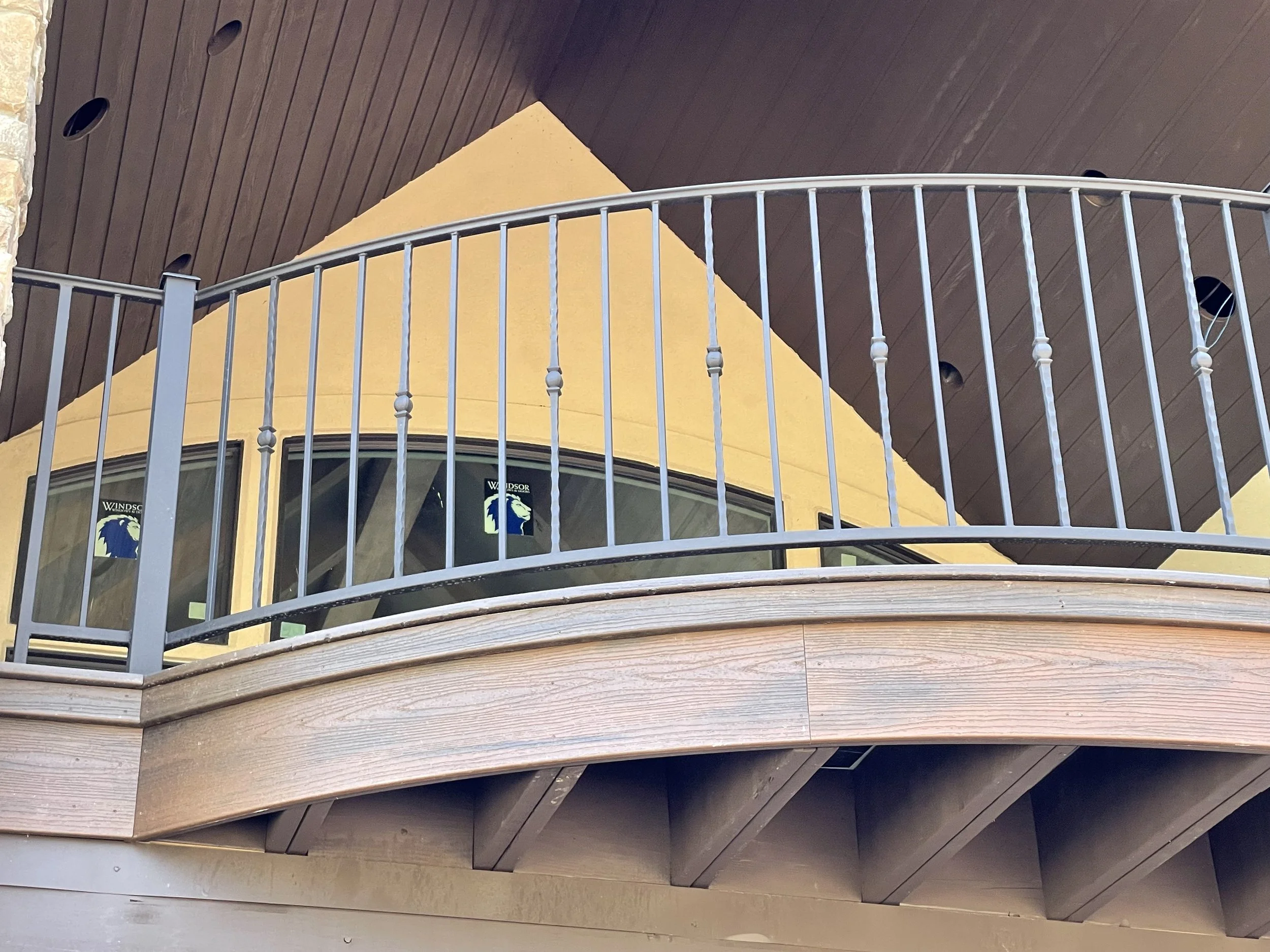 Multiple curved wooden shelves with a metal railing in the foreground, part of a building with a yellow wall and dark brown ceiling, and a window with a logo reading 'WINDSOR' and a lion. Circular recessed lights on the ceiling.