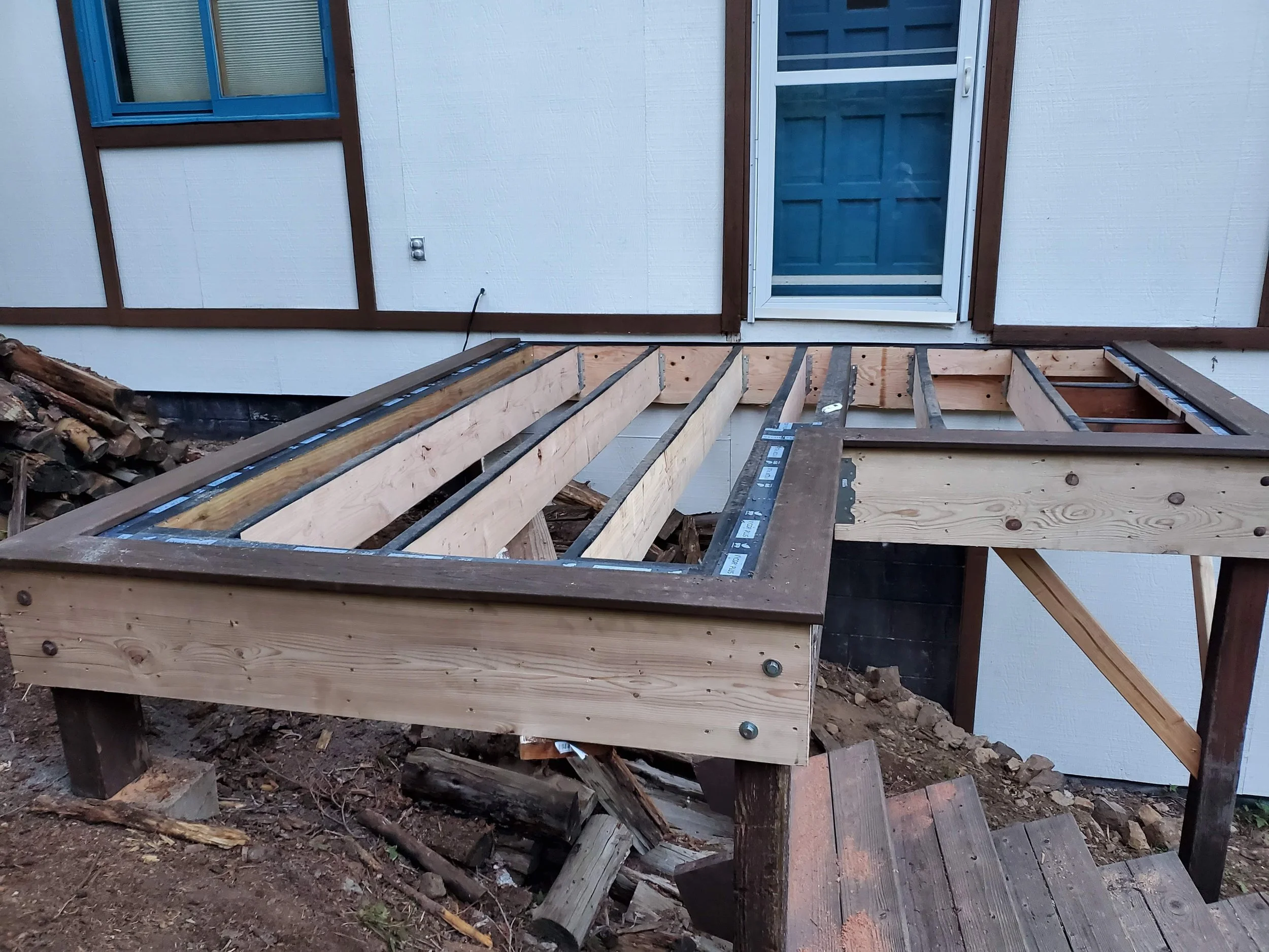 A house with white siding, a blue window, and a blue storm door, with a wooden deck or porch under construction in the foreground, including wood framing and tools.