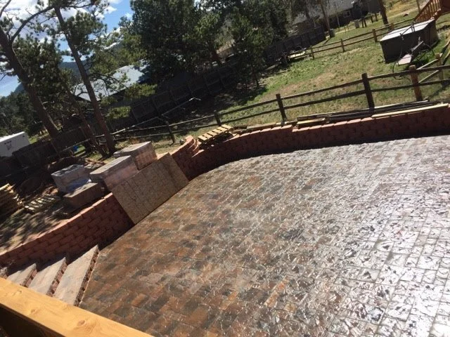 A backyard with a newly constructed brick patio surrounded by a wooden fence, with some outdoor furniture and trees in the background.
