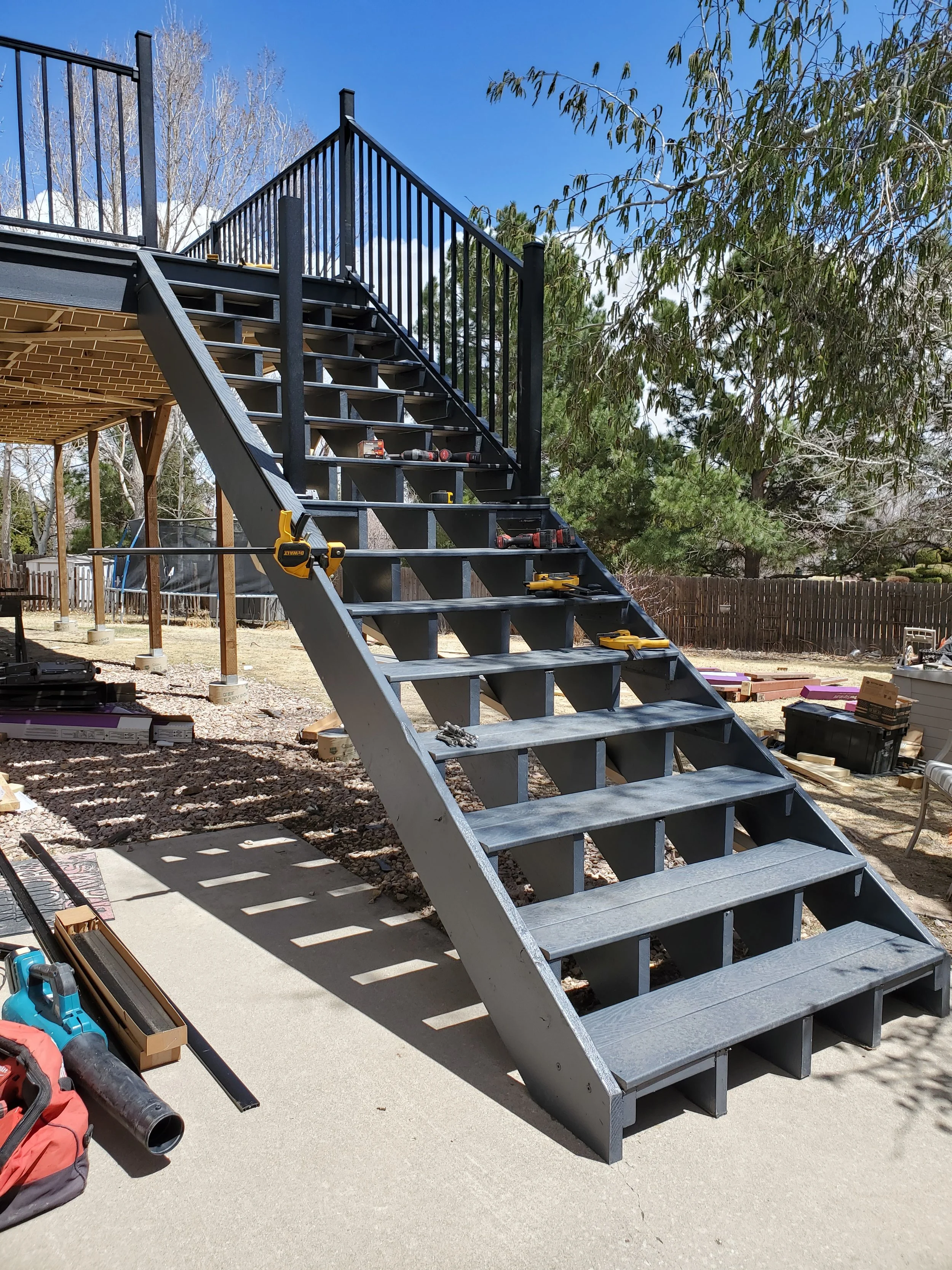 An outdoor construction site with a stairway made of metal, partly assembled, with tools and supplies on and around it. There is a backyard with trees and a wooden fence.
