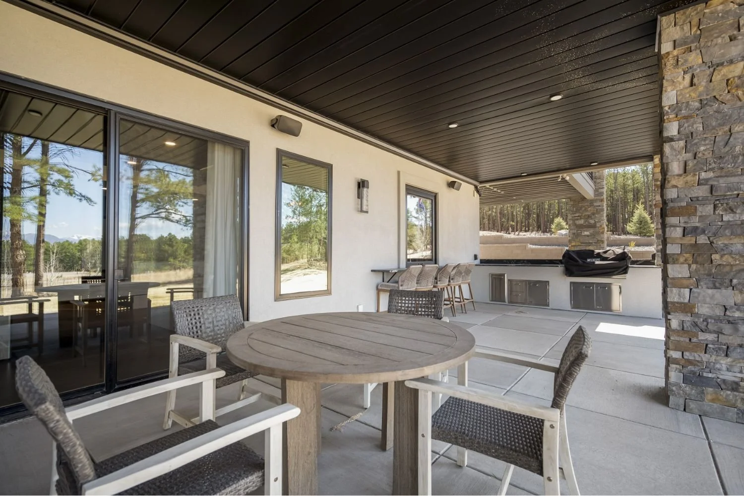 Outdoor patio with a wooden table and chairs, a counter with barstools, and a built-in grill, surrounded by trees and a stone pillar.
